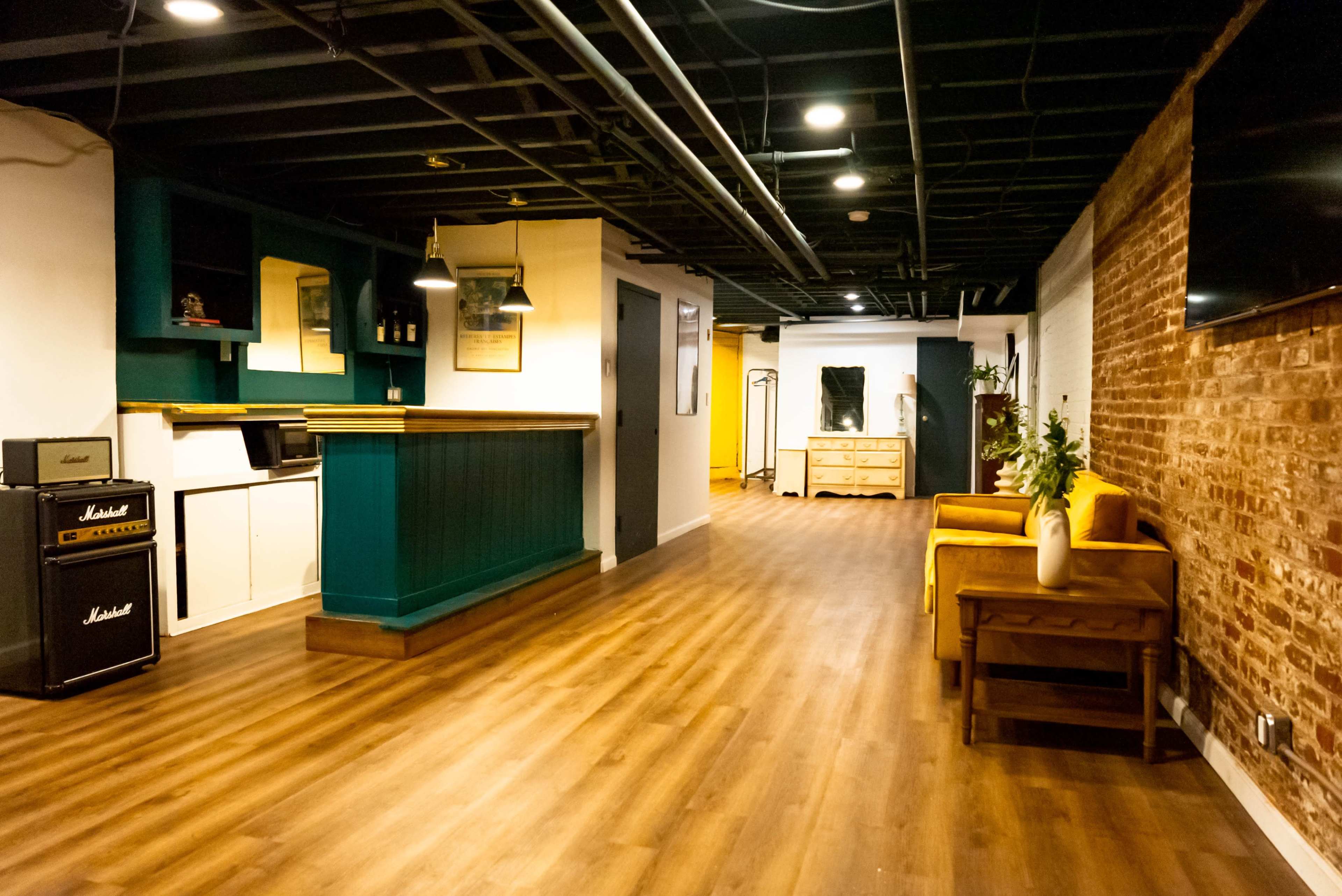A spacious indoor area featuring a combination of hardwood flooring, teal accent walls, and exposed brick, with furniture including a yellow sofa and a black amplifier.