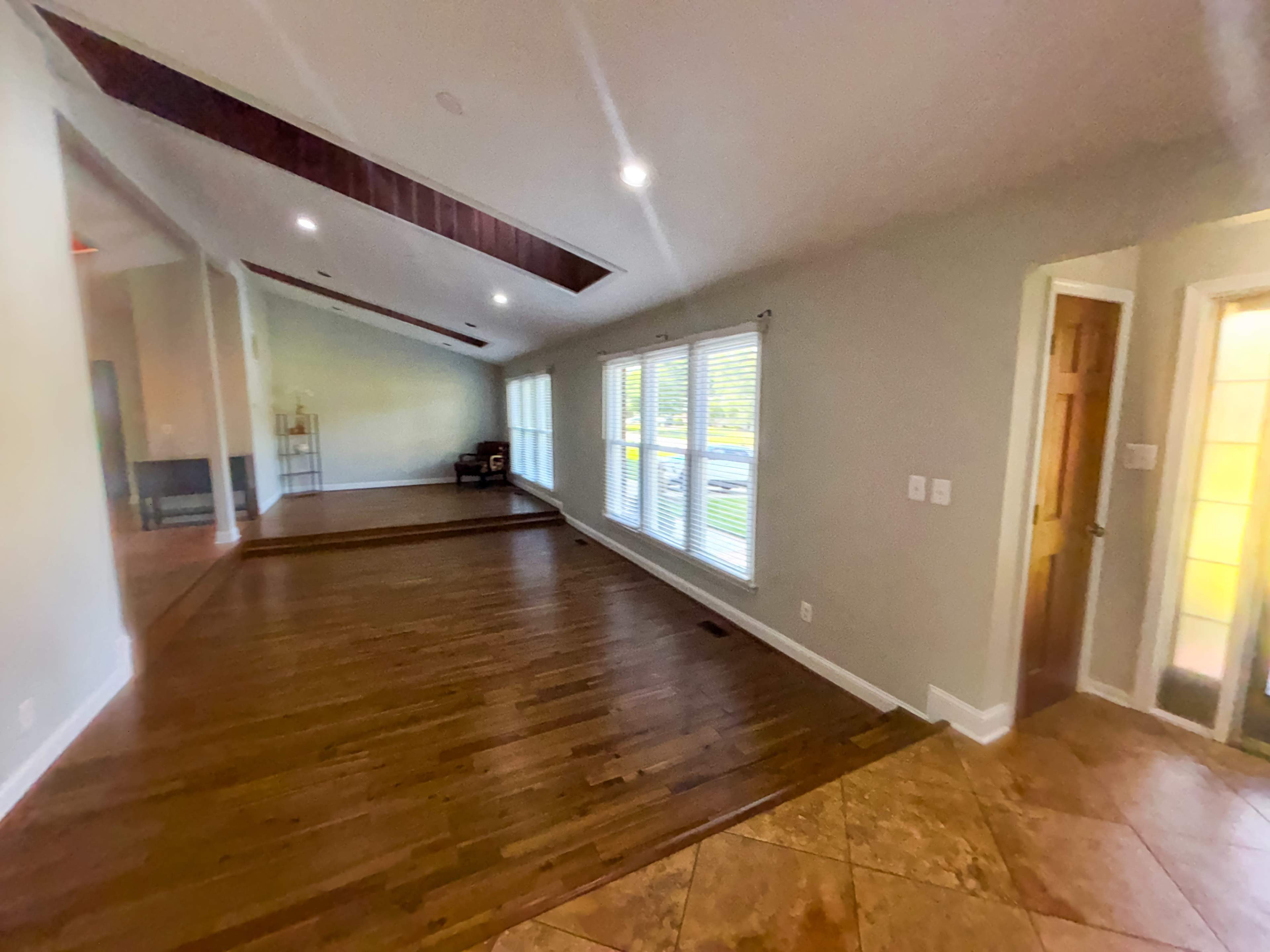 The image shows a spacious room with hardwood flooring, large windows allowing natural light, and a doorway leading to another area.