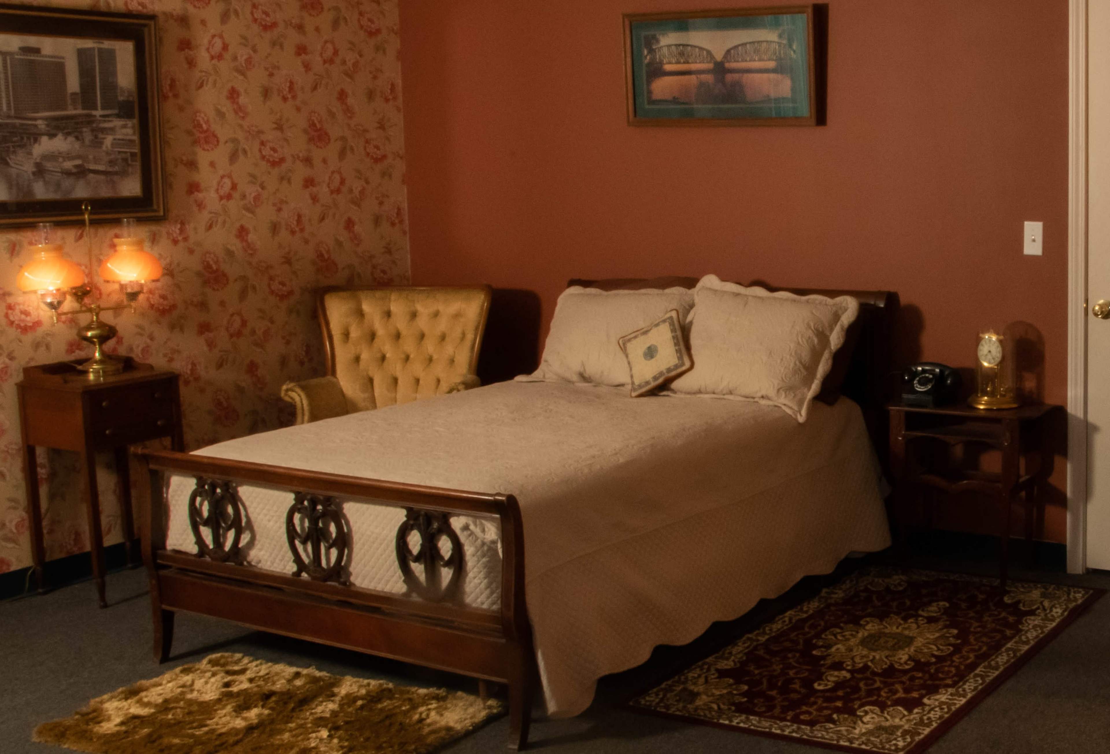A vintage-style bedroom featuring a wooden bed with a white bedspread, a yellow upholstered chair, and a side table with a lamp and a telephone.