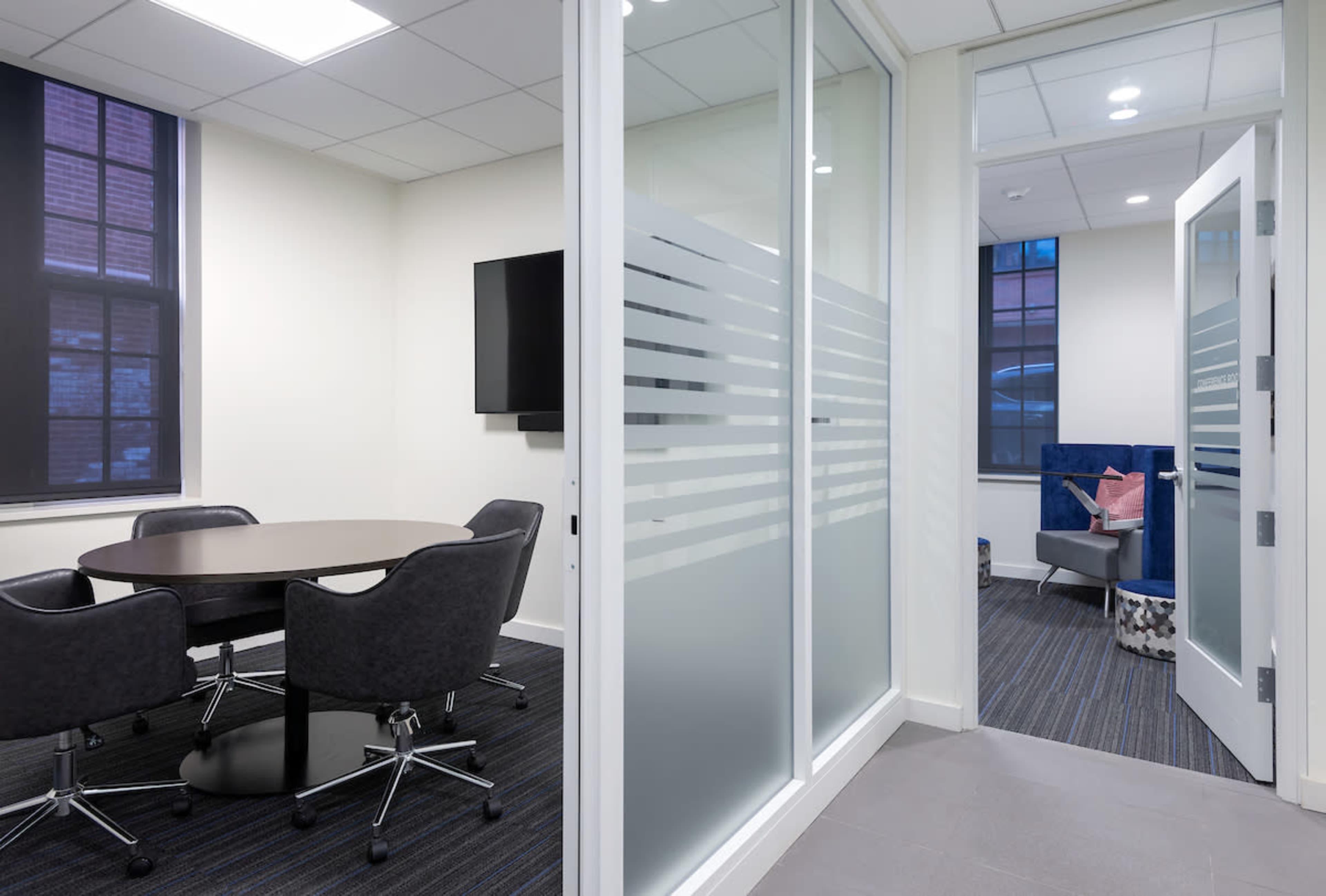 A modern office space features a conference room with a round table and chairs, adjacent to a seating area visible through a glass door.