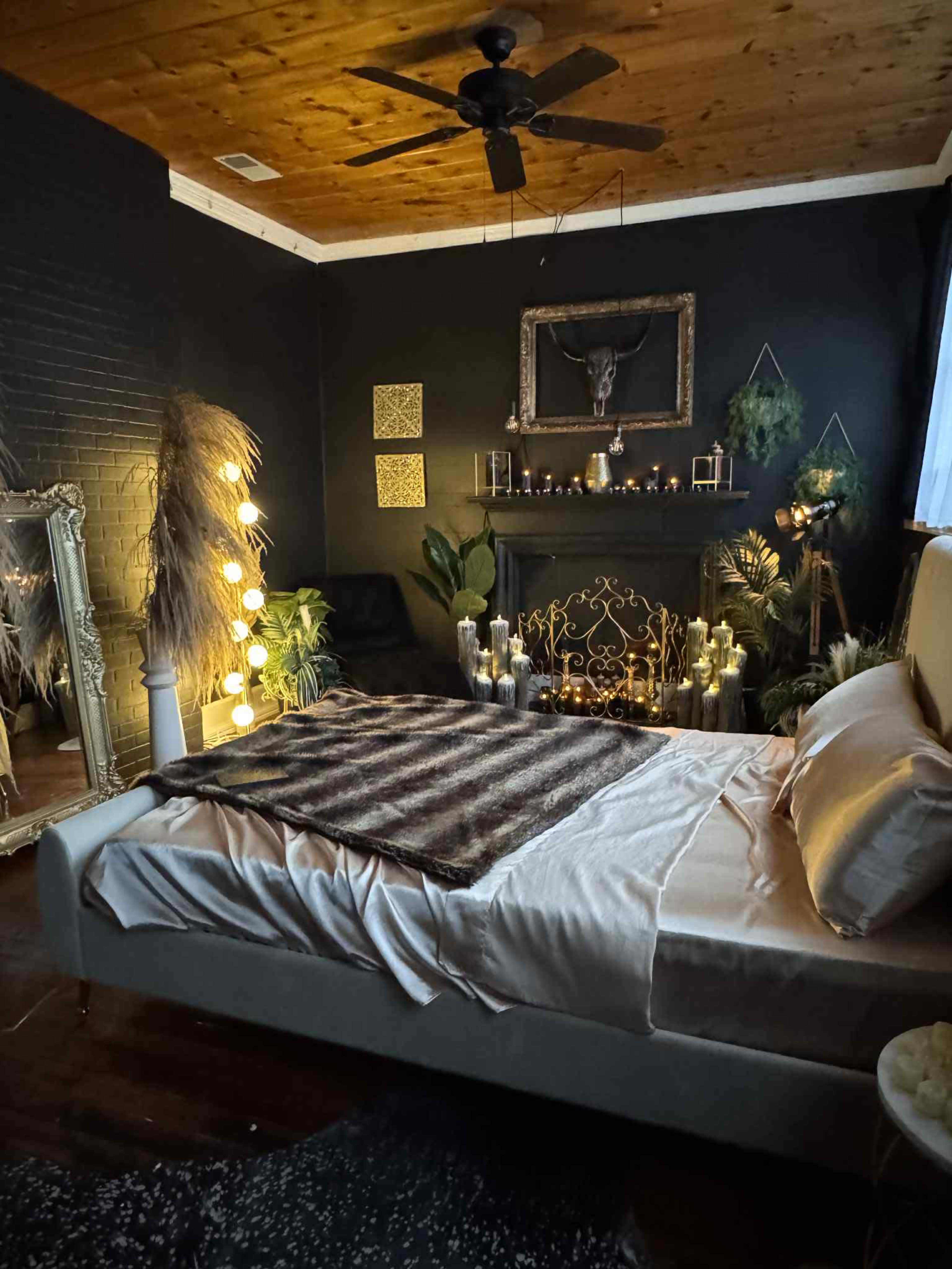The image shows a dark-themed bedroom with a large bed, decorative plants, and ambient lighting, featuring candles and a mirror on one side.