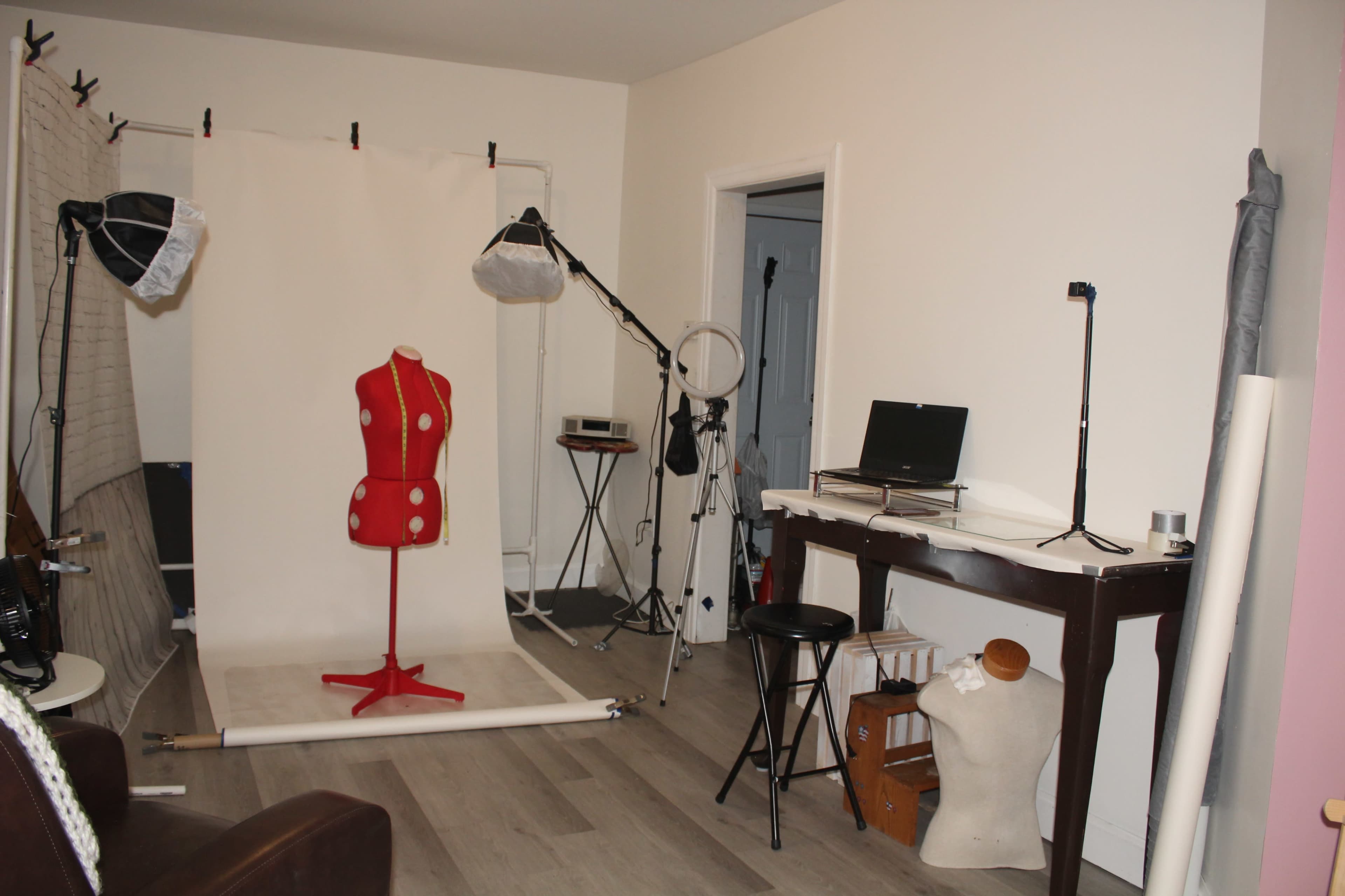 The image shows a small photo studio with a dress form, lighting equipment, a desk with a computer, and various props arranged against a white backdrop.