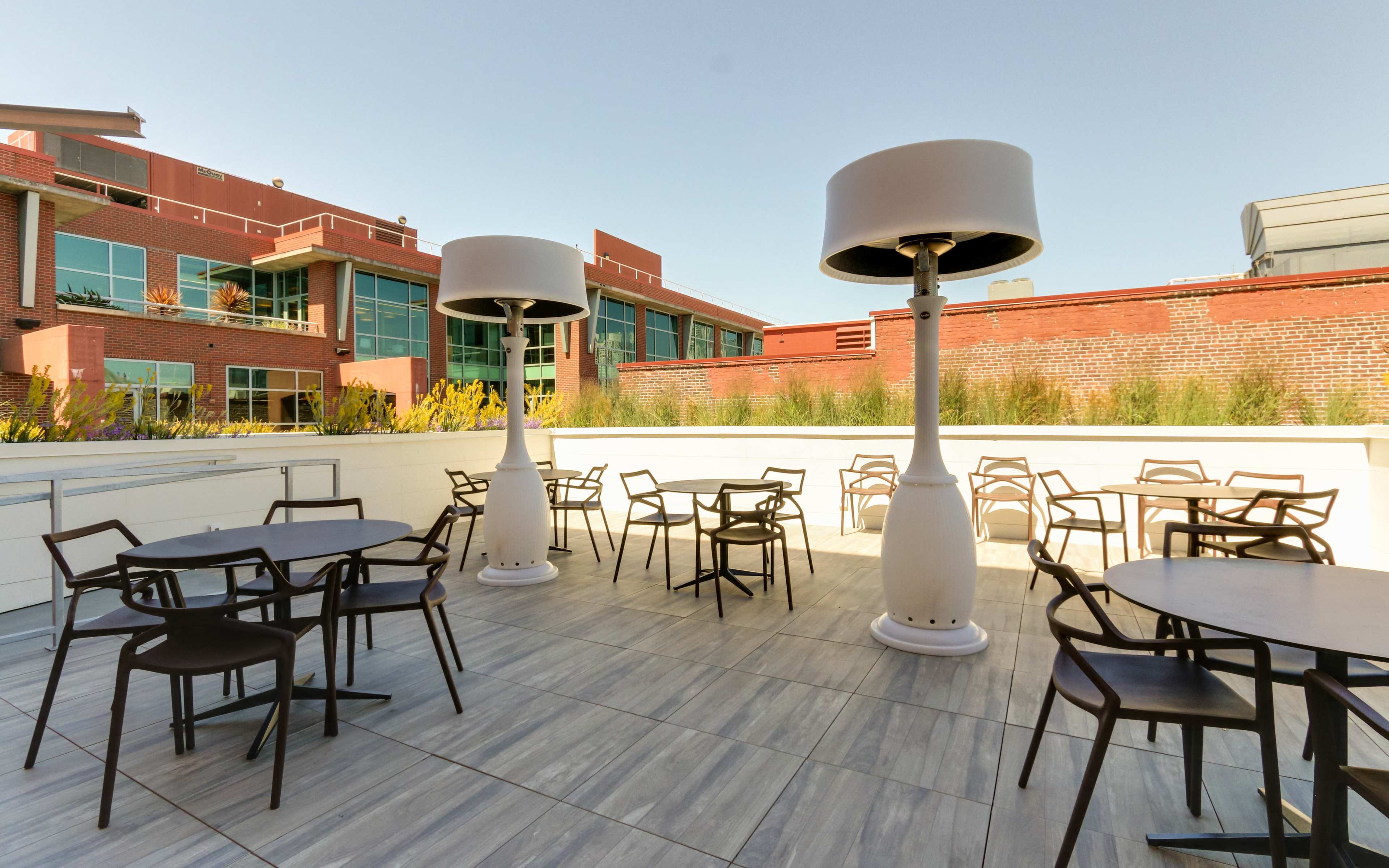 An outdoor patio area with several tables and chairs, two large heaters, and buildings in the background.