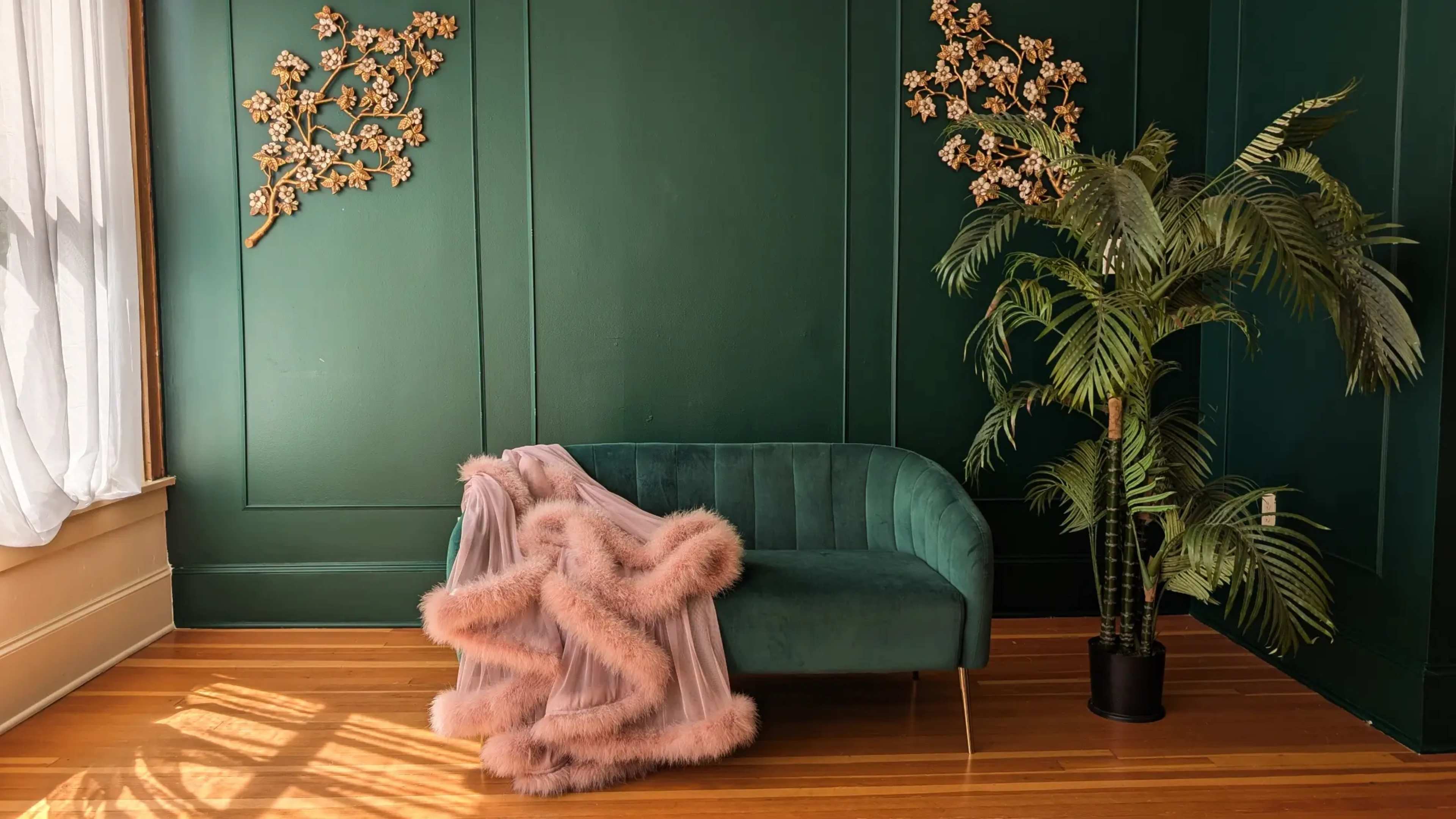 A green-walled room features a velvet sofa draped with a pink throw and a potted plant in the corner.