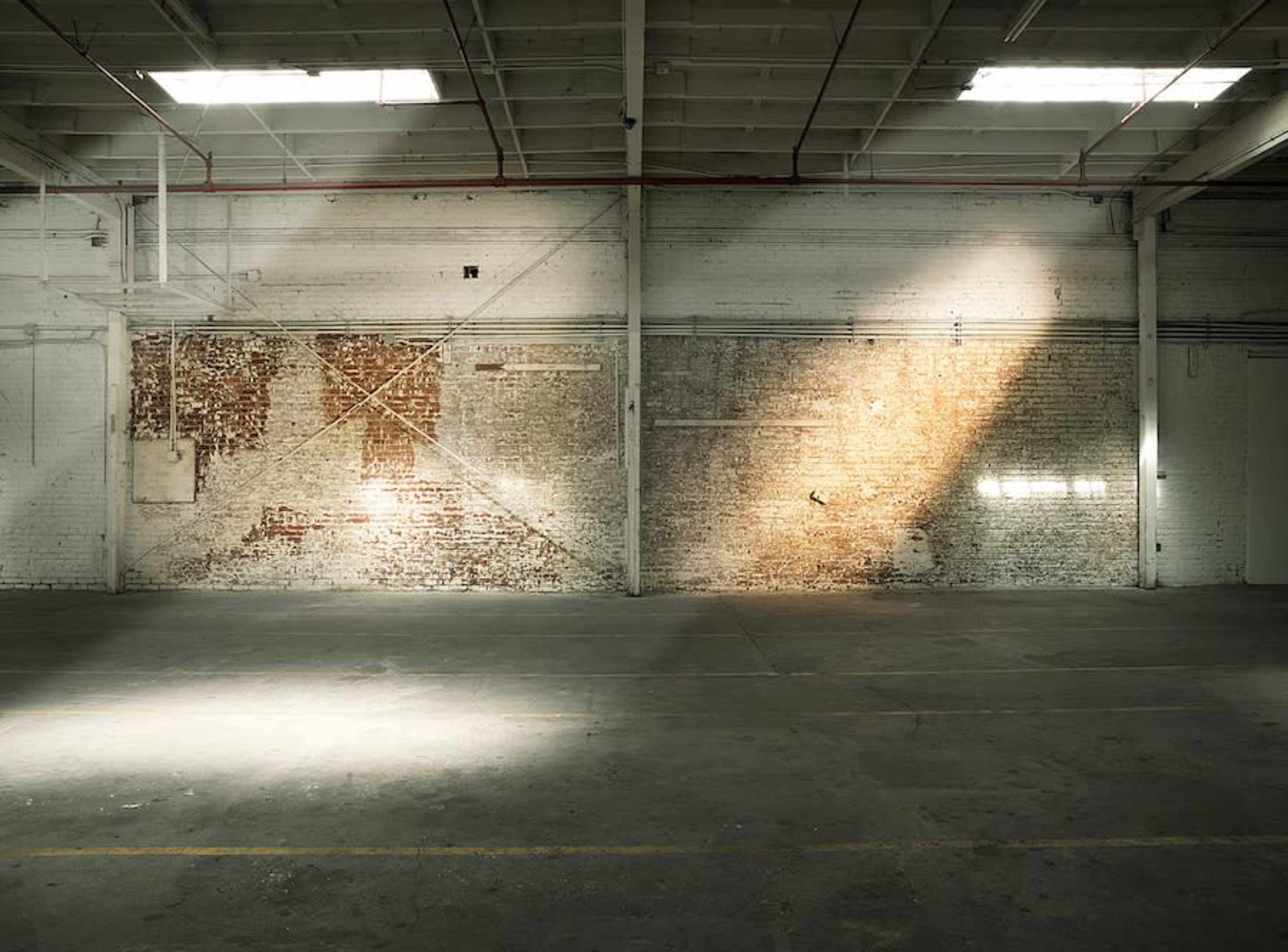 The image shows a spacious, empty warehouse with exposed brick walls and a beam of sunlight illuminating a section of the floor.