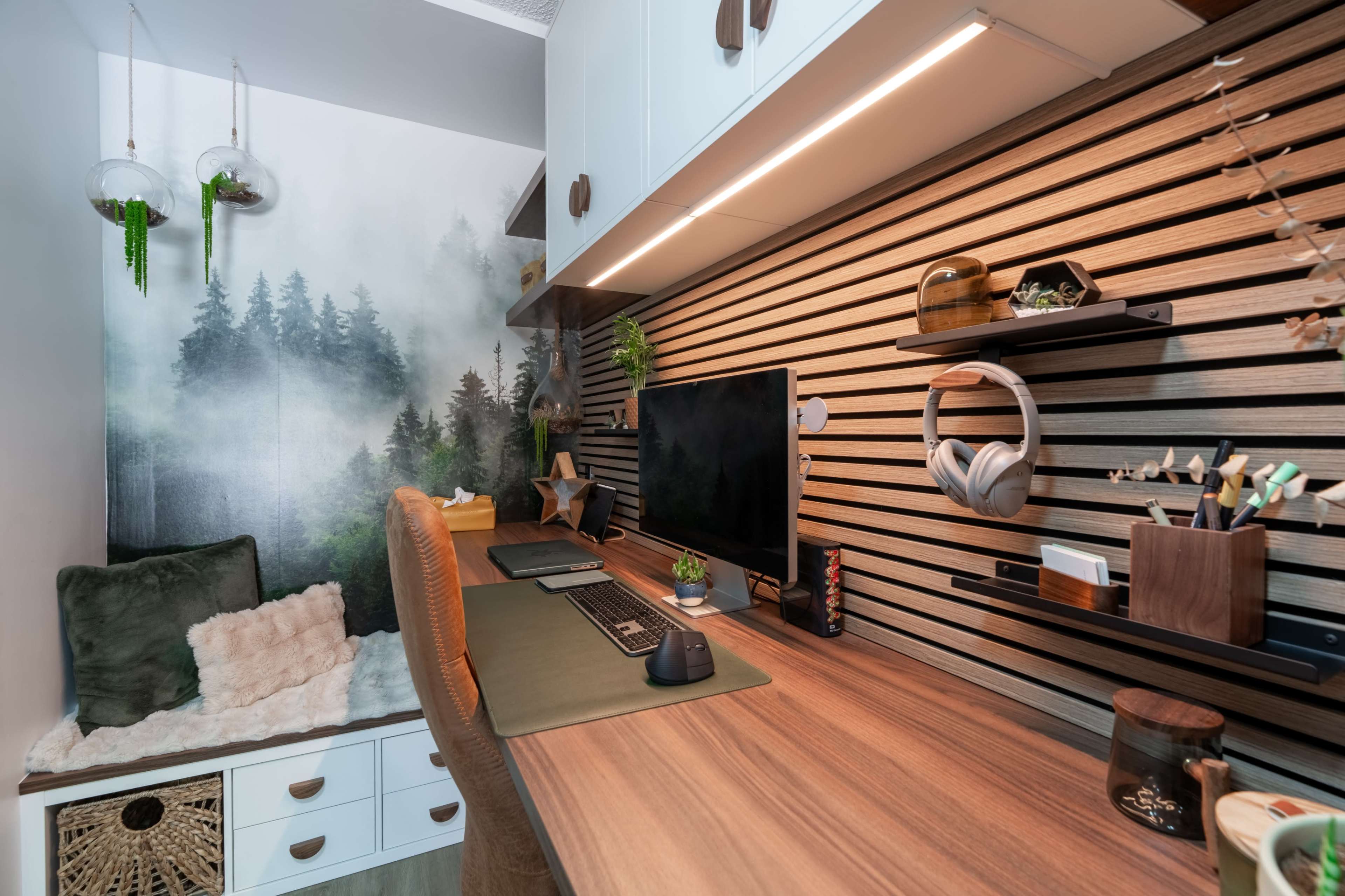 The image shows a modern workspace with a wooden desk, a computer monitor, and organized accessories against a backdrop of forest-themed wallpaper and shelves.