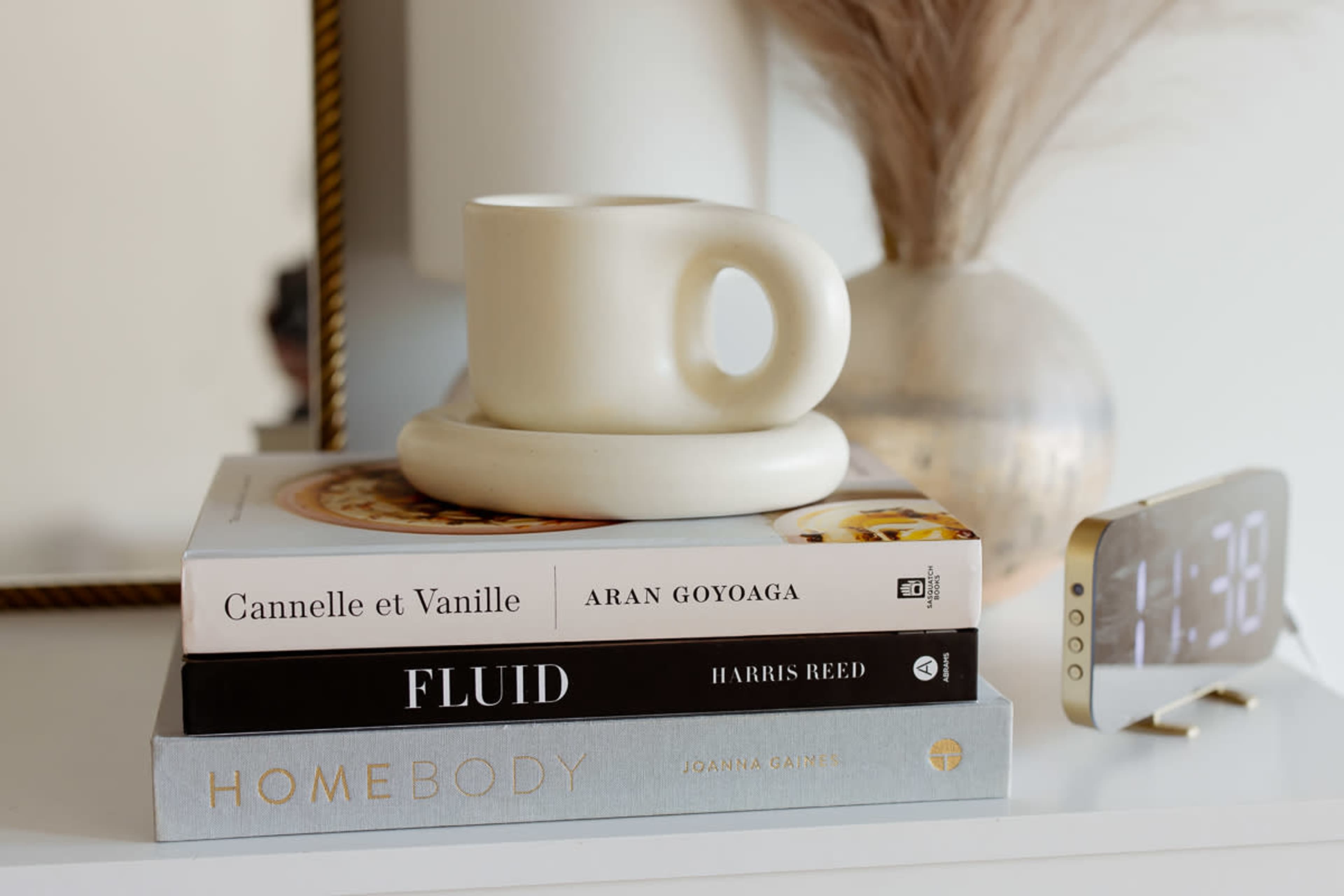 A stacked arrangement of books and a decorative mug is placed on a surface next to a digital clock showing the time as 11:38.