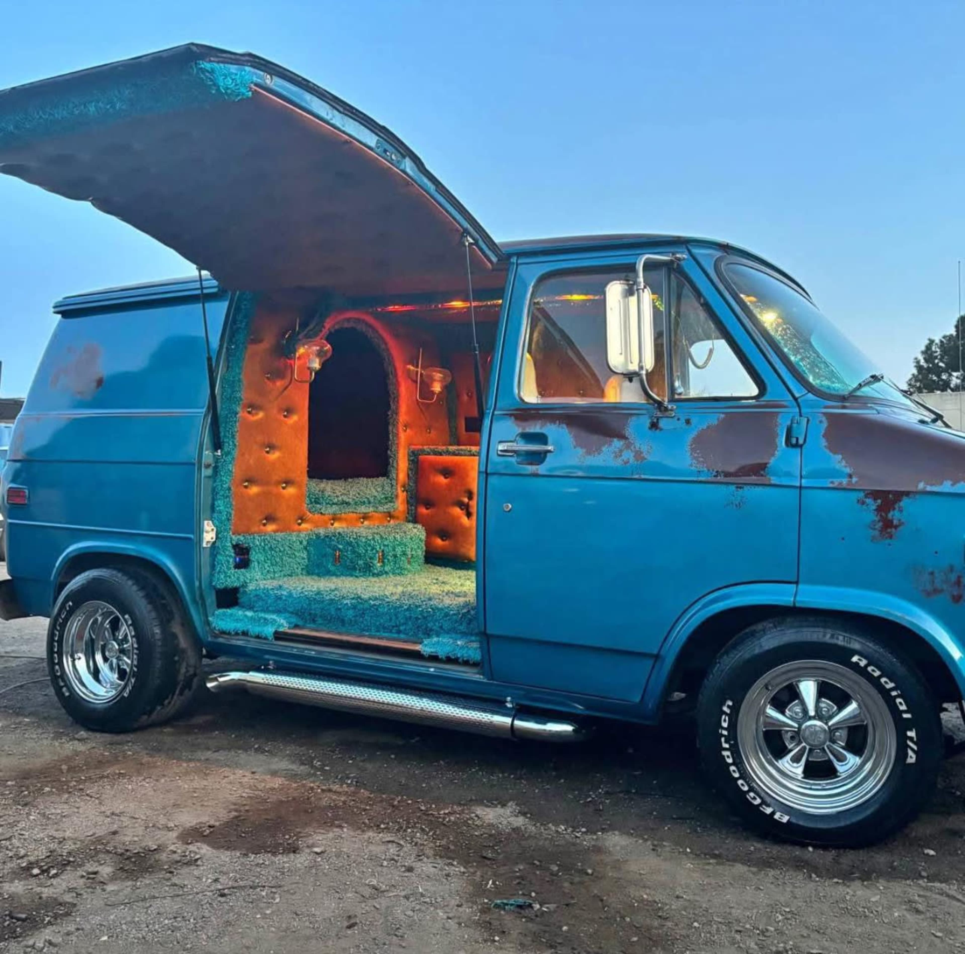 A vintage blue van with a rusted exterior and an open door reveals a colorful, carpeted interior featuring a rounded alcove.