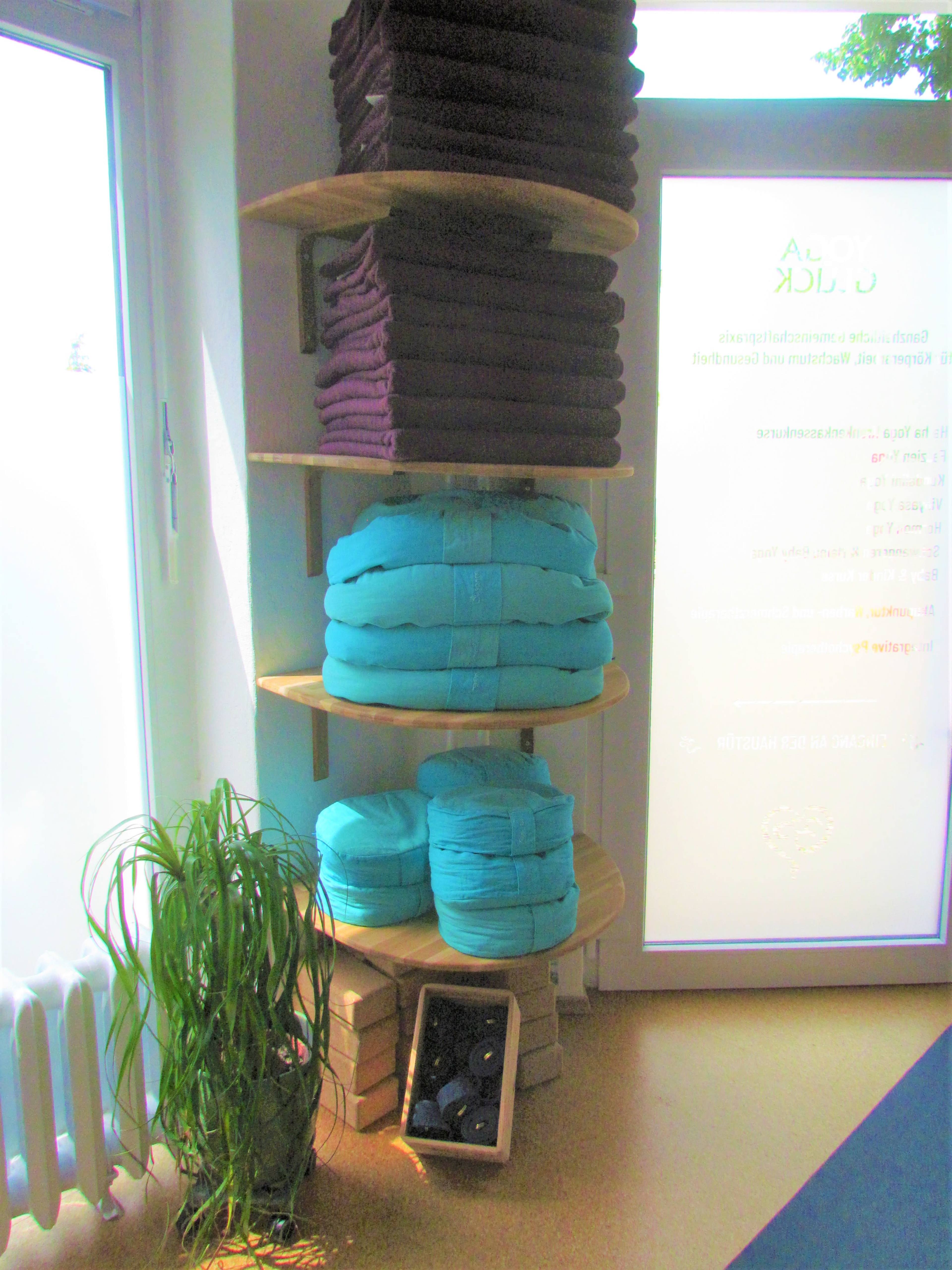 A corner of a room featuring neatly stacked towels in shades of turquoise and purple on wooden shelves, with a potted plant beside them and a partially visible window displaying information.