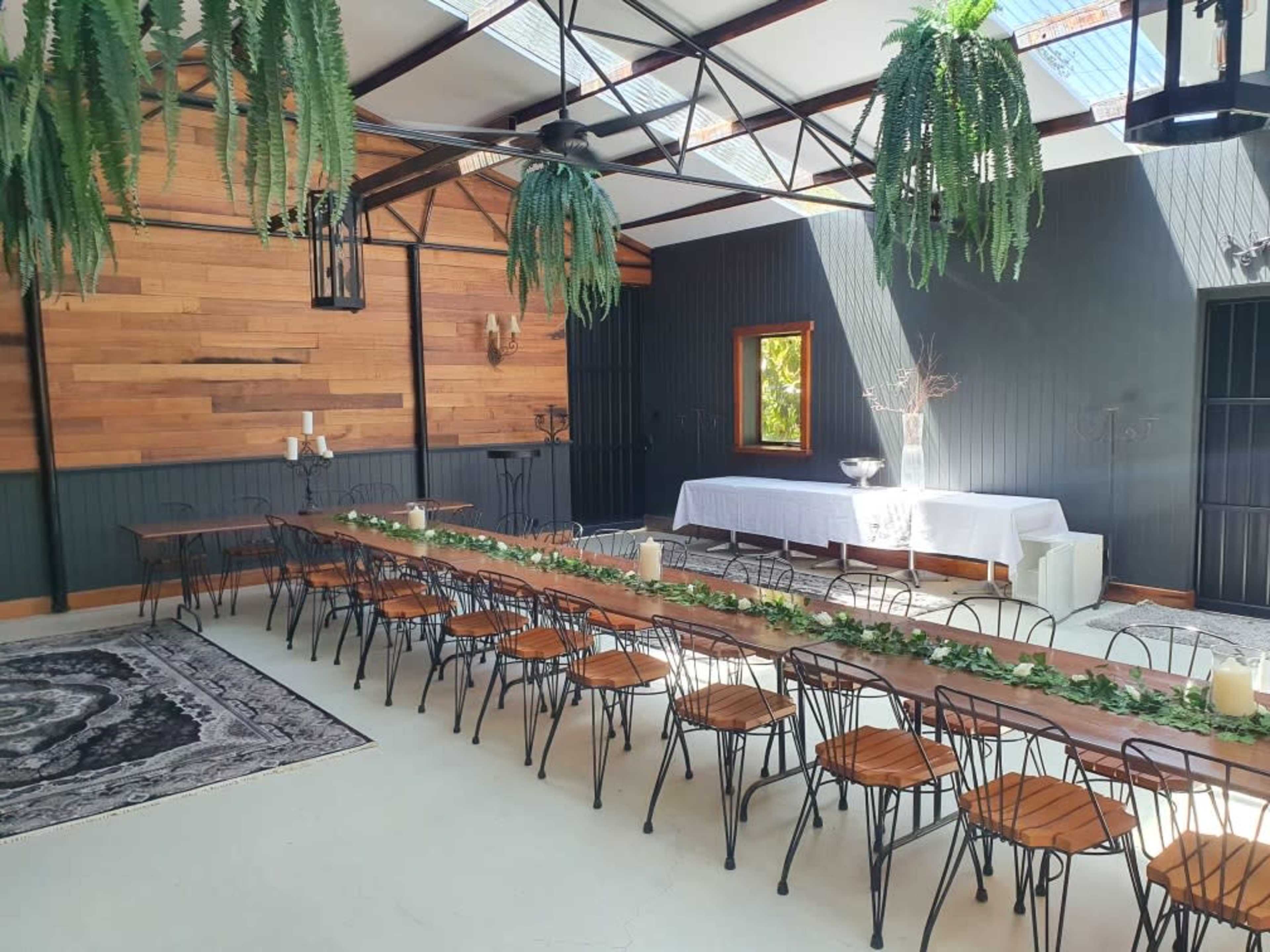 A spacious indoor venue features long wooden tables with black chairs, adorned with greenery, against a backdrop of wooden paneling and large windows.