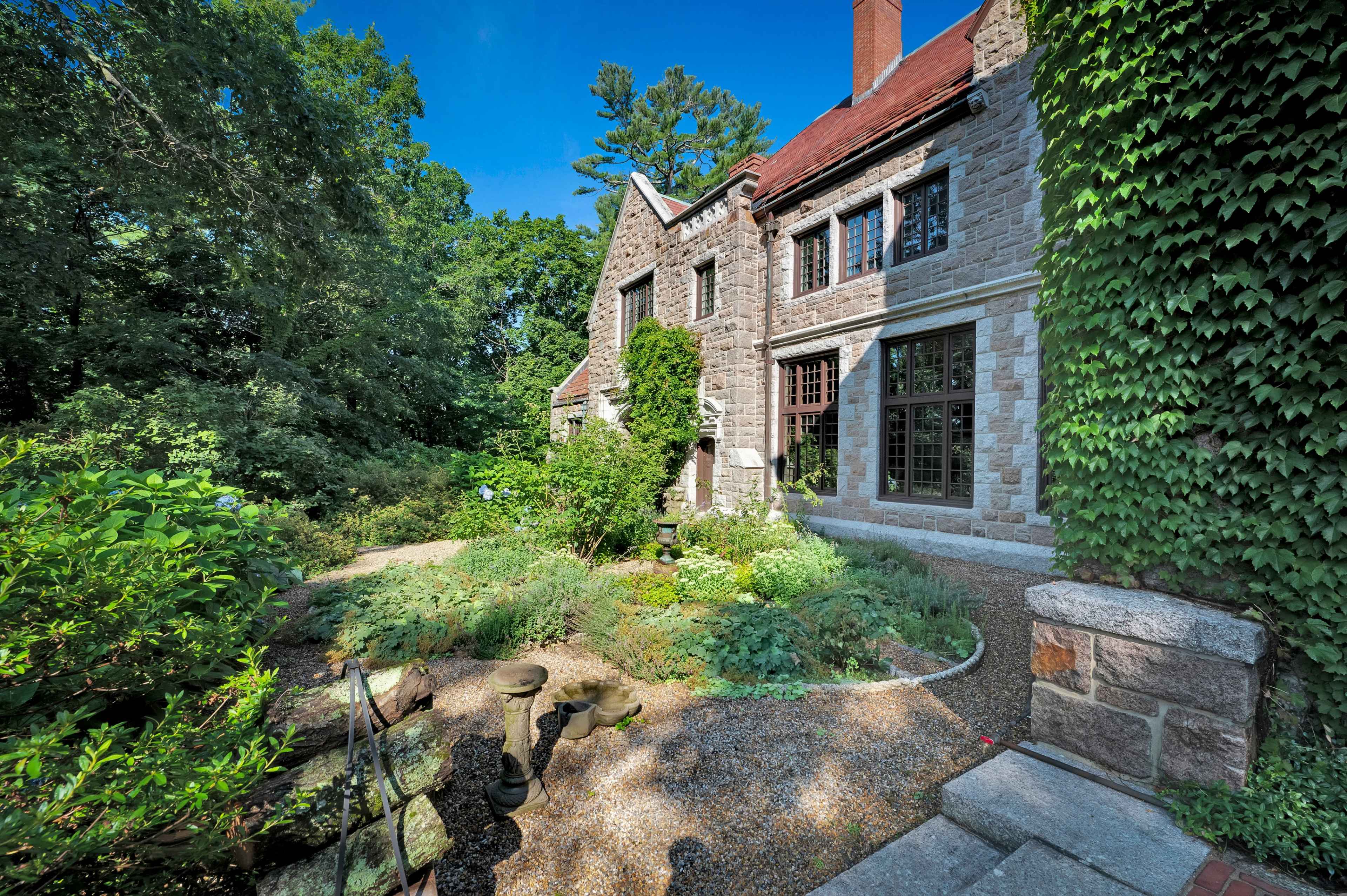 A stone house with a red roof is surrounded by lush greenery and a garden pathway.
