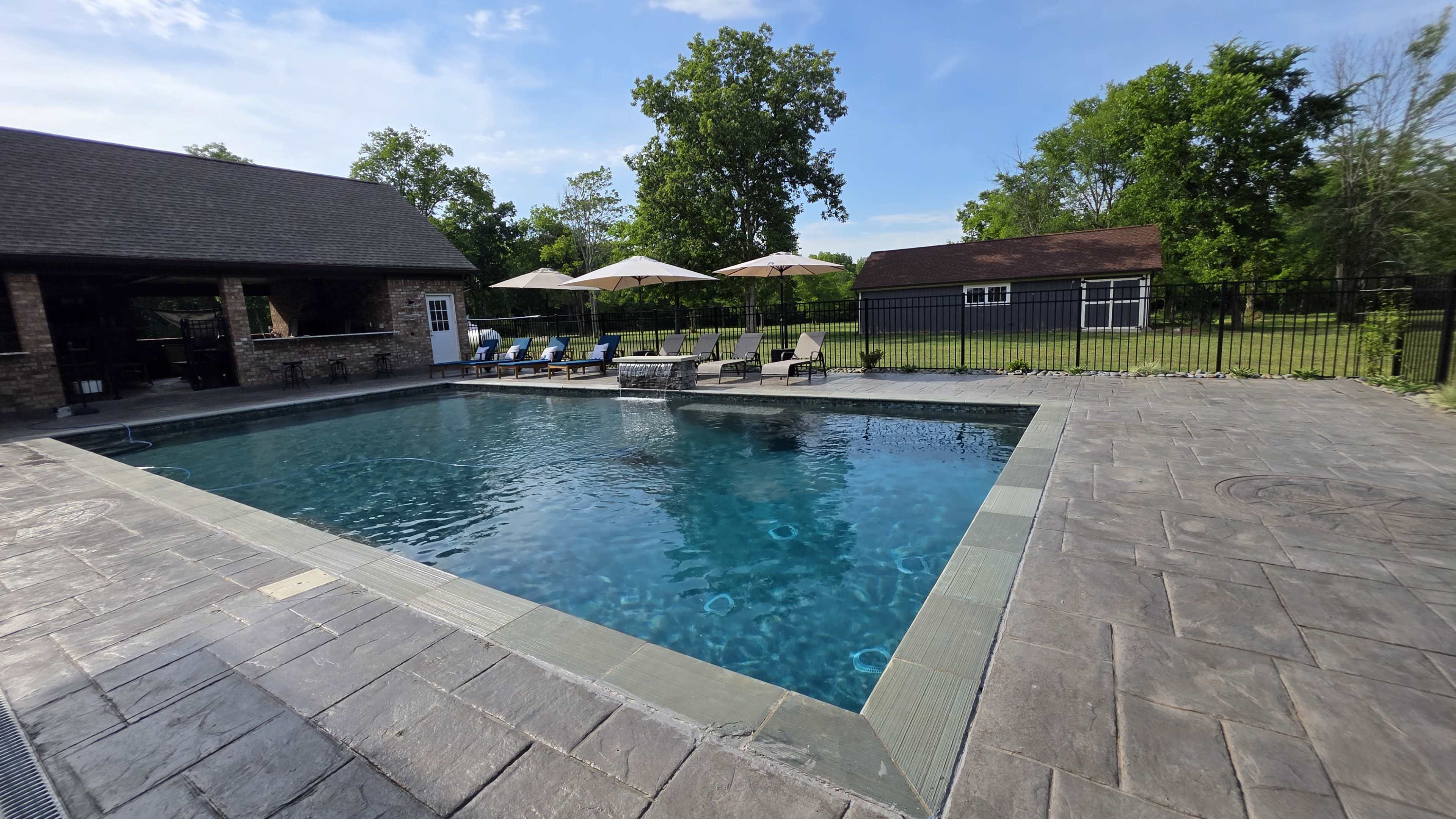 The image shows a rectangular swimming pool surrounded by a patio with lounge chairs and umbrellas, set in a grassy backyard with trees and a shed in the background.