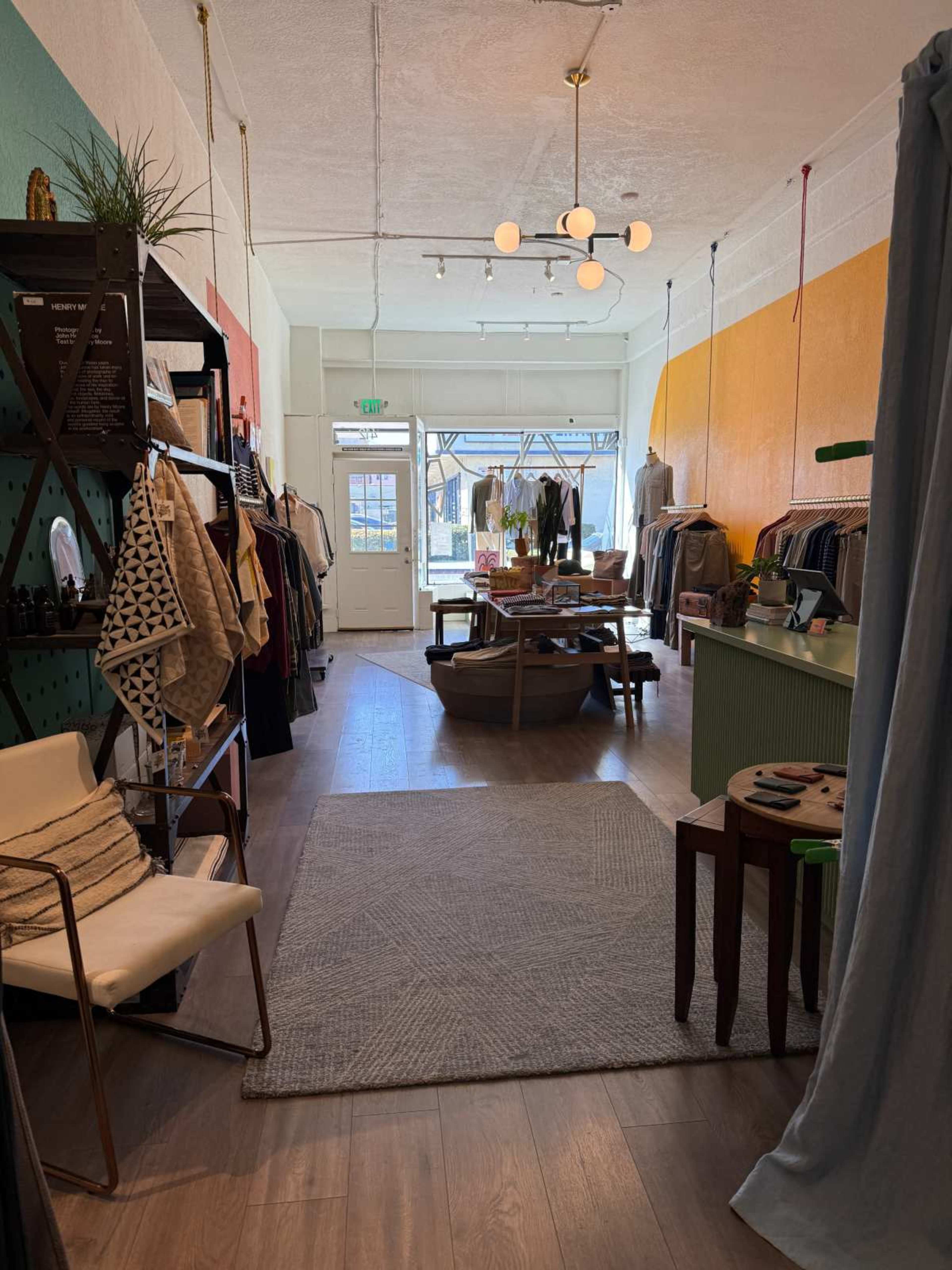 The image shows a boutique interior featuring clothing racks, shelves with decorative items, and a seating area, all illuminated by natural light coming through large front windows.