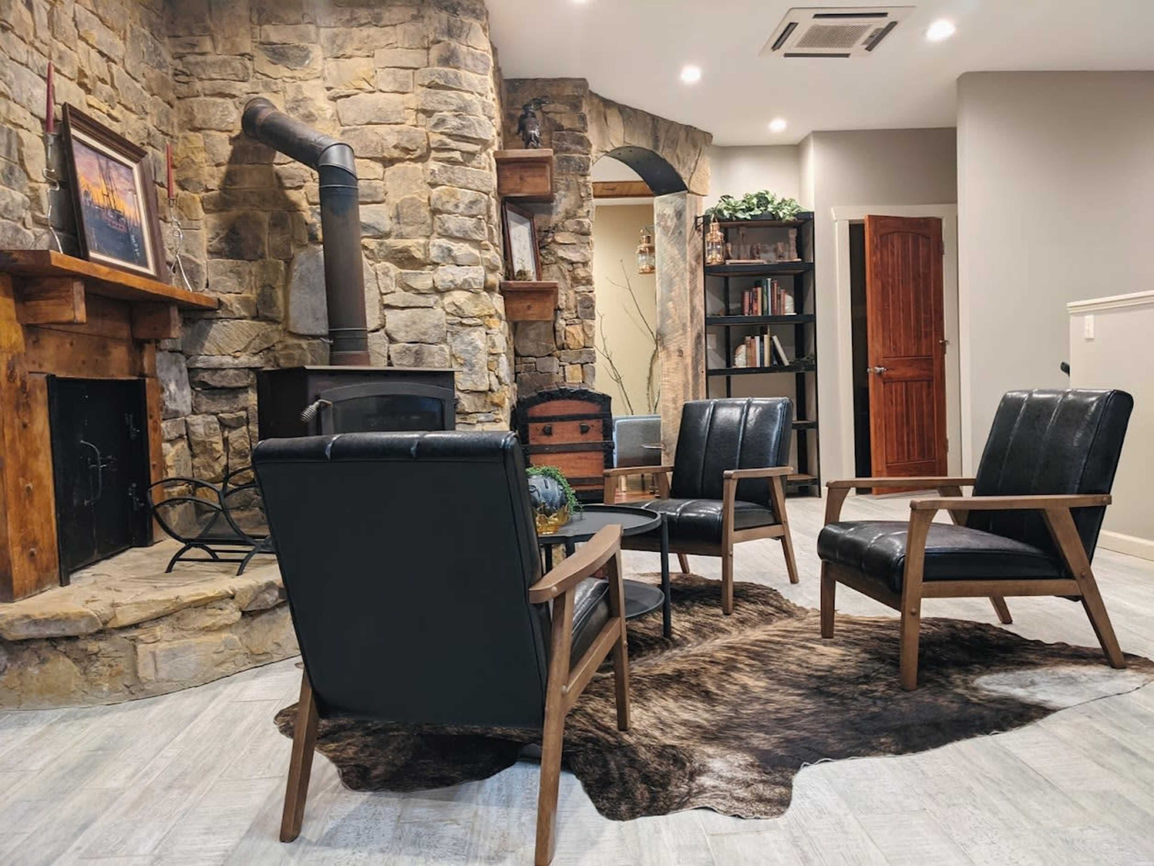 A cozy living area with a stone fireplace, three black leather armchairs arranged on a faux fur rug, and bookshelves in the background.