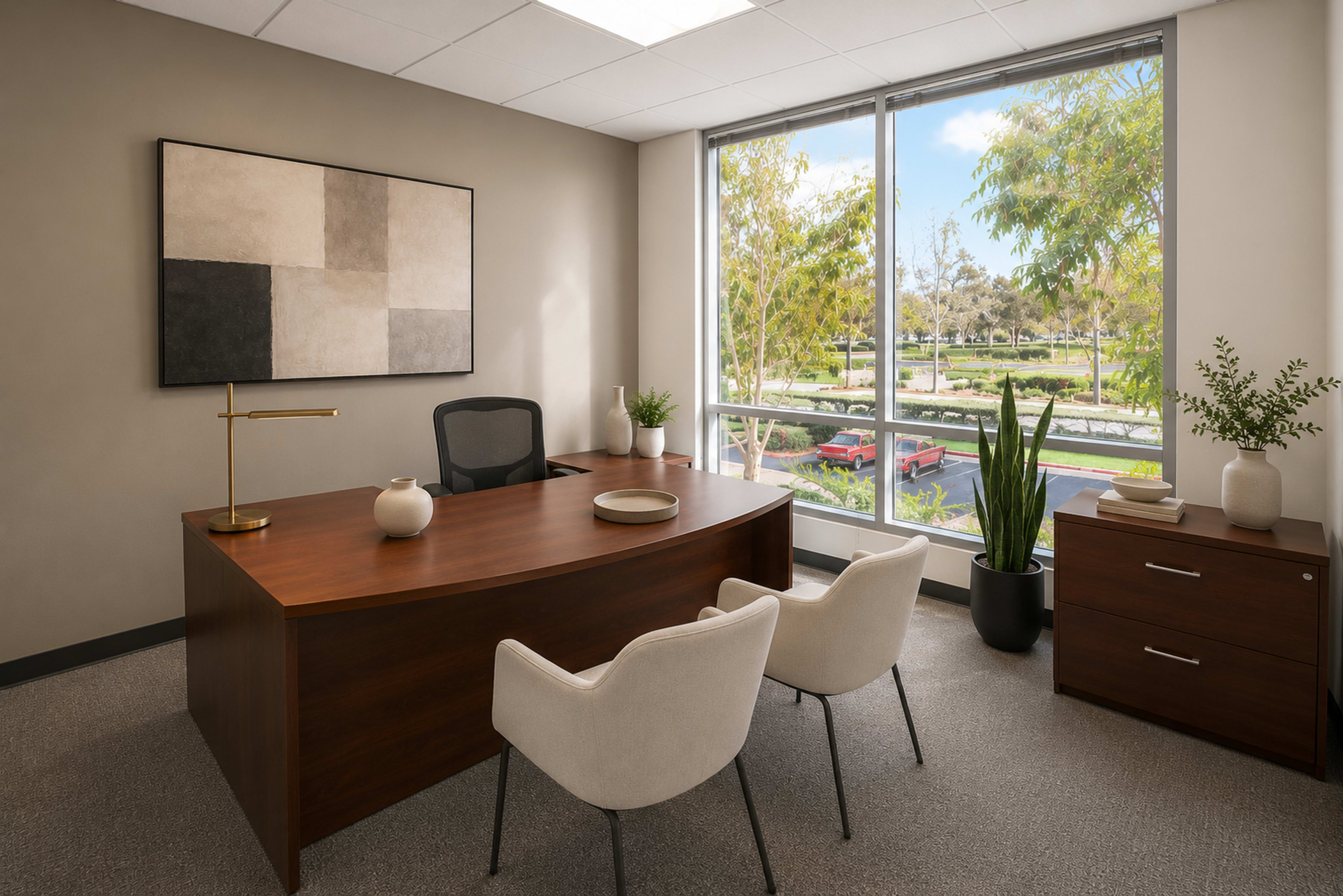 The image shows a modern office space with a wooden desk and two white chairs, featuring large windows that overlook a green area and parked cars outside.
