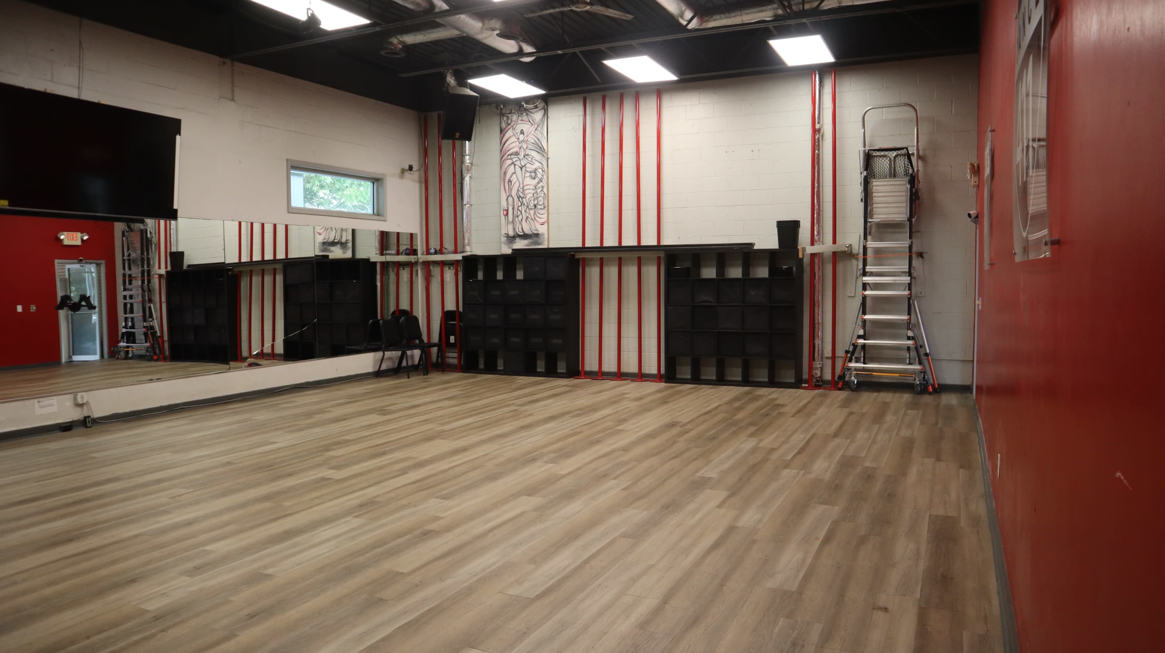 The image shows an empty dance studio with wooden flooring, mirrored walls, and a red accent wall, featuring a shelving unit and a ladder in the background.