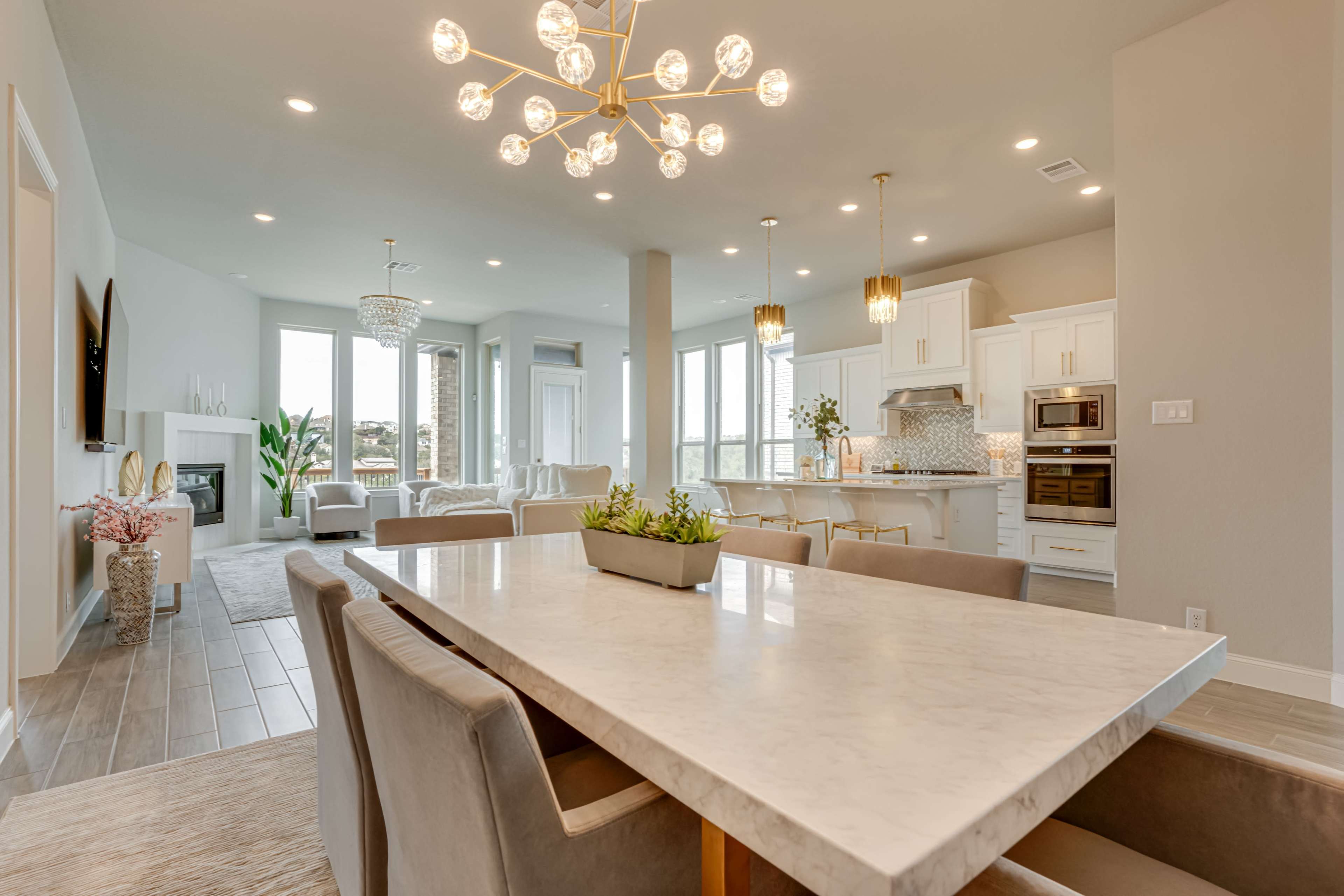 A modern dining area with a marble table, surrounded by upholstered chairs, leading into an open-concept kitchen and living space filled with large windows.
