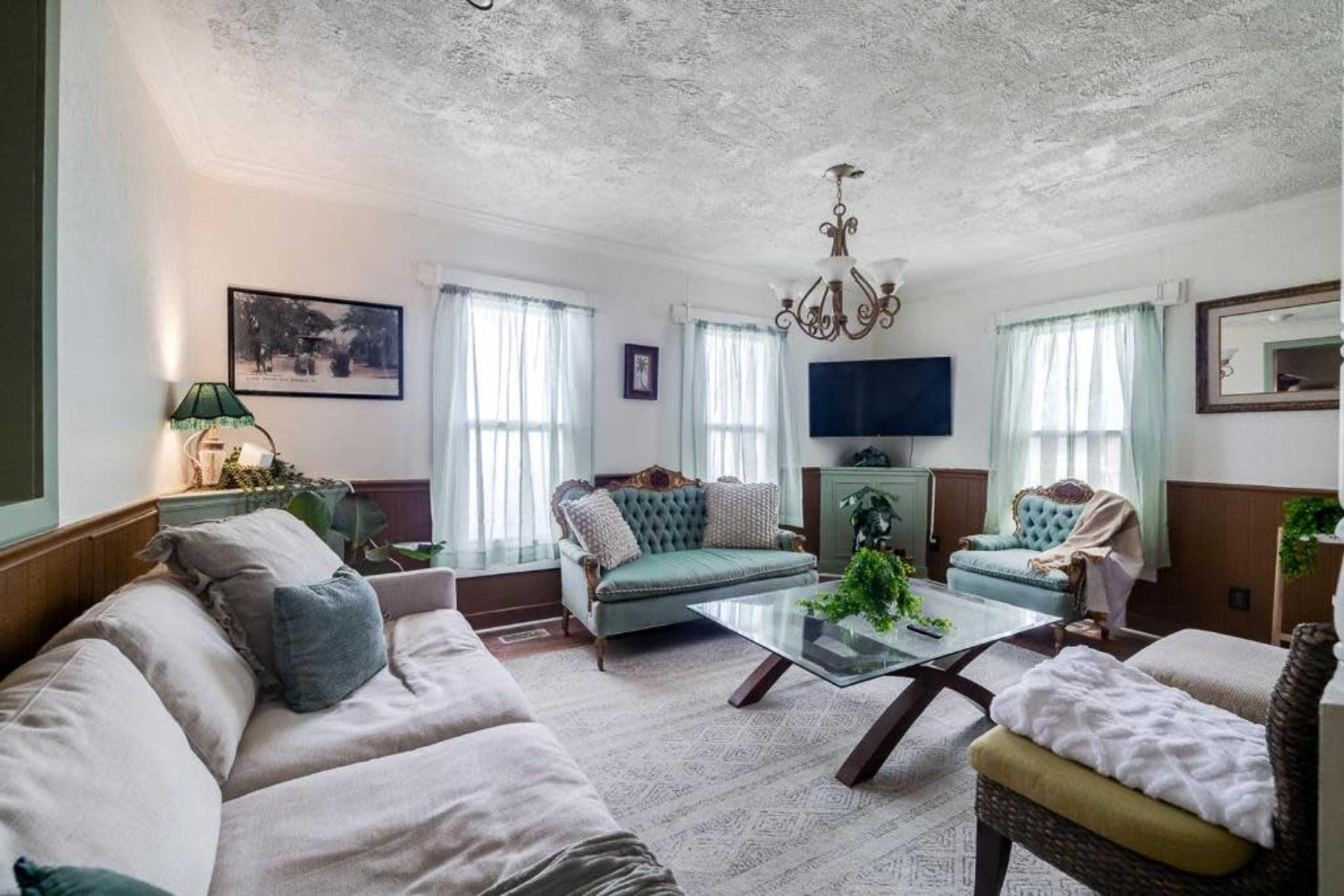 A living room with two sofas, a glass coffee table, a television mounted on the wall, and large windows with sheer curtains.