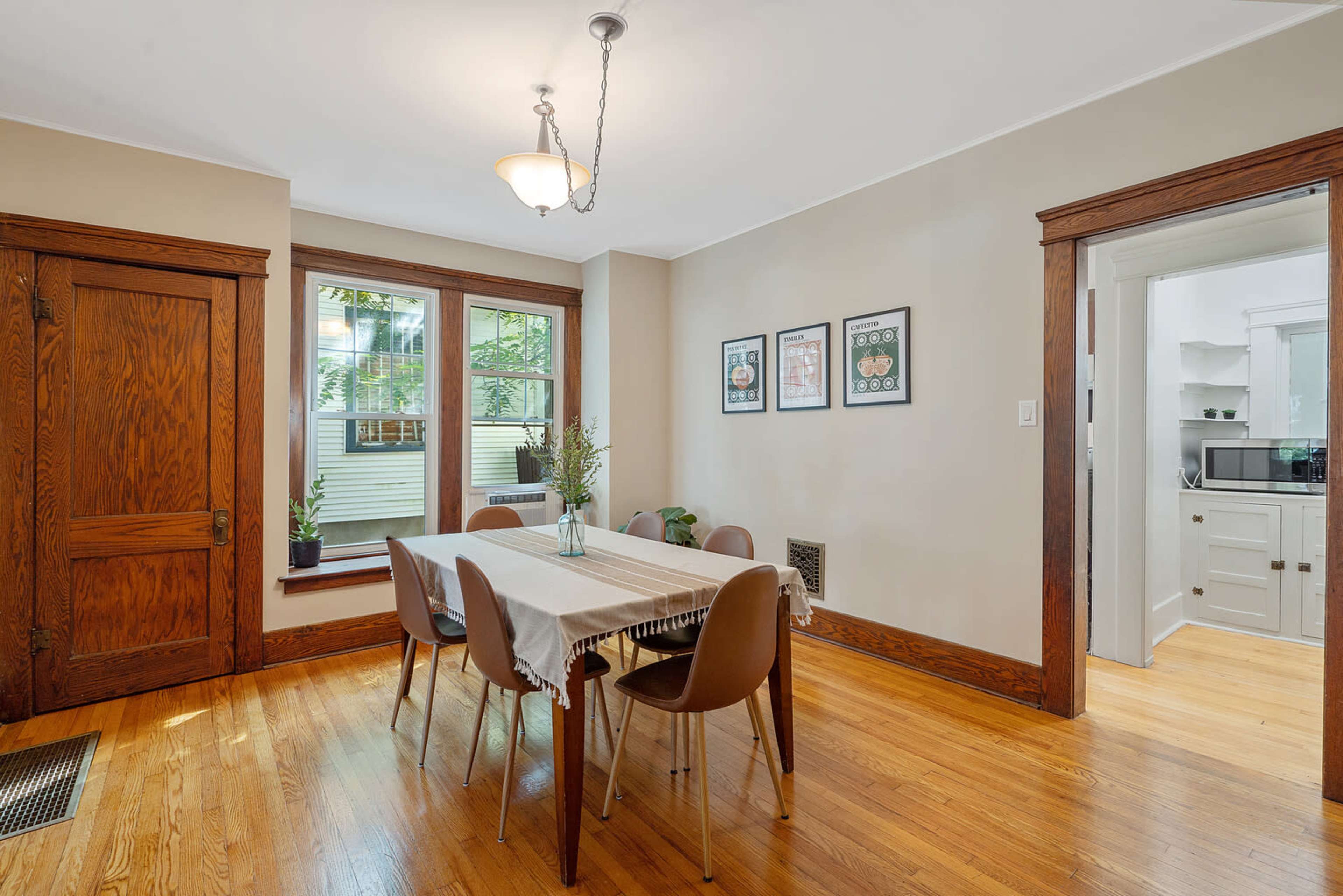 A dining room features a wooden table with chairs, large windows, and adjacent access to a kitchen.