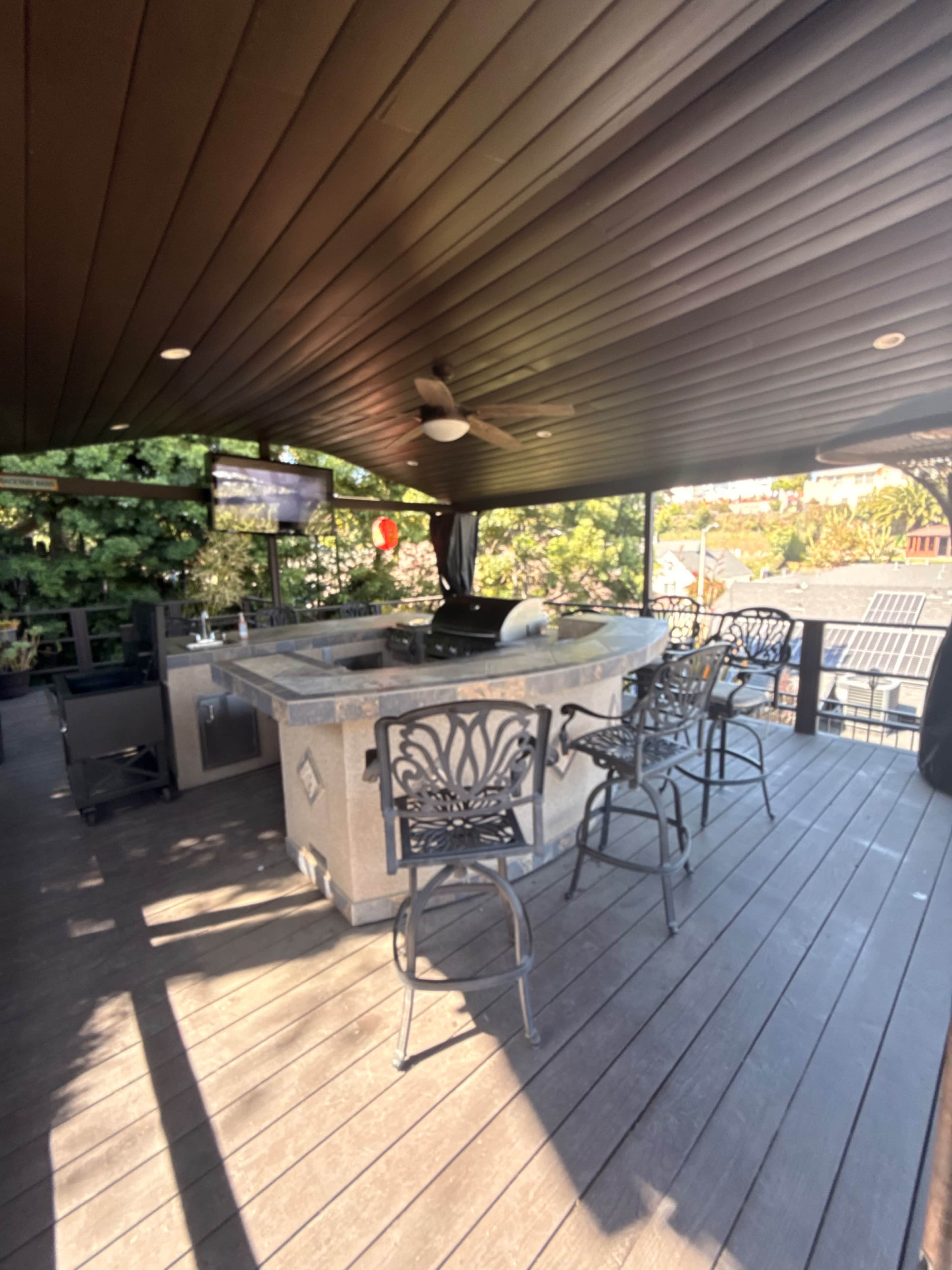 The image shows an outdoor bar area with a stone counter and several black metal stools, set under a covered wooden structure.