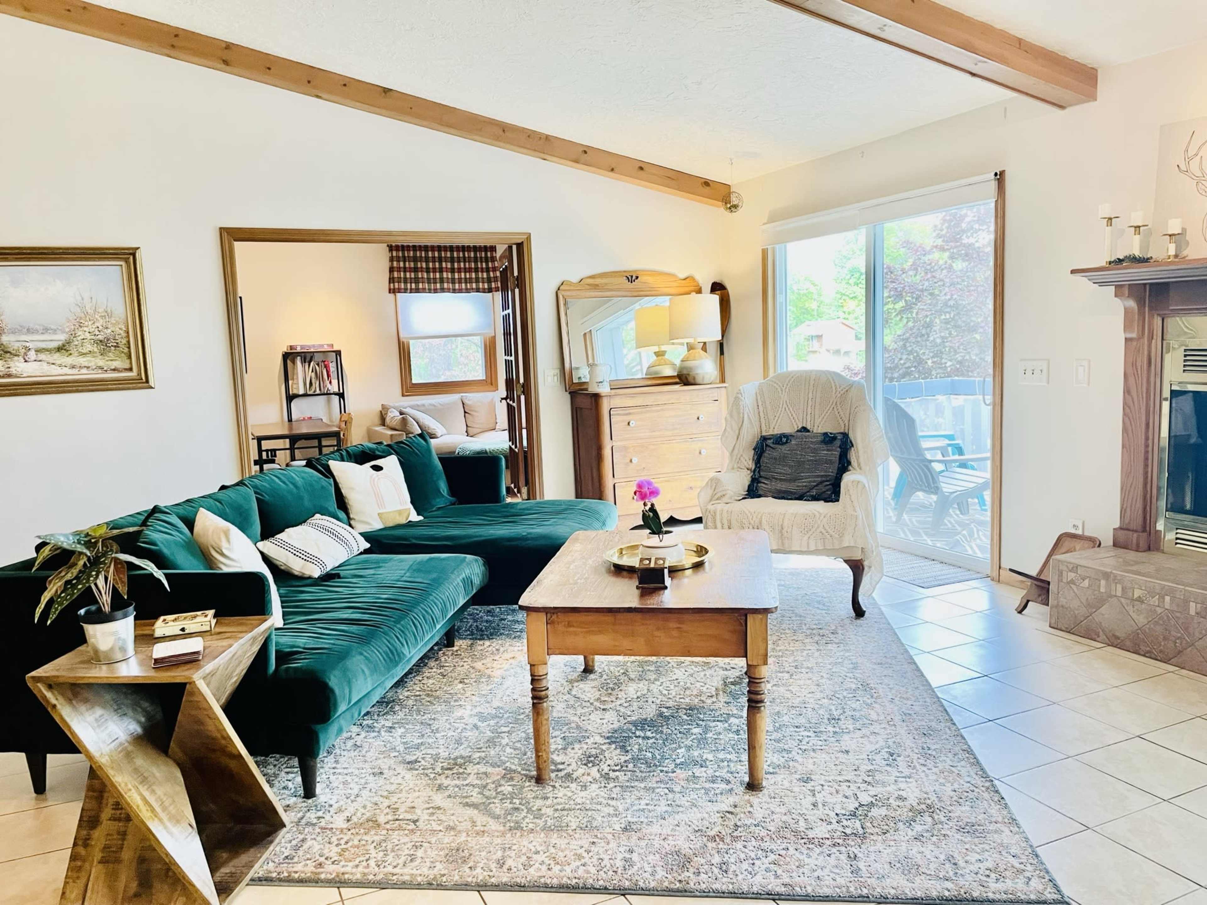 The room features a green sectional sofa, a wooden coffee table with flowers, and a light-colored chair, all arranged around a fireplace and large windows that allow natural light to enter.
