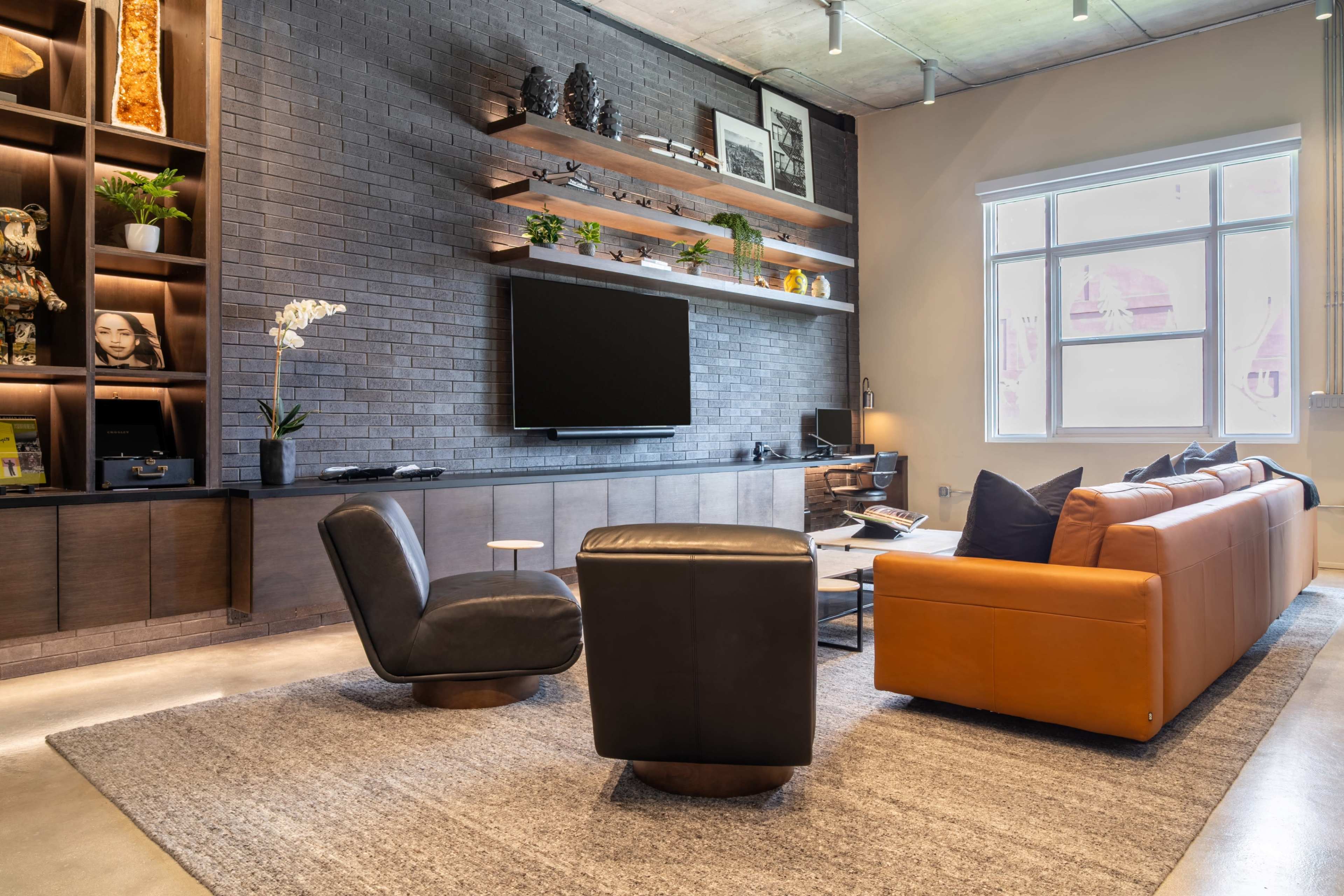 The image shows a modern living room with a dark brick wall, wooden shelves, a television, and a combination of a black armchair and an orange sofa on a large area rug.