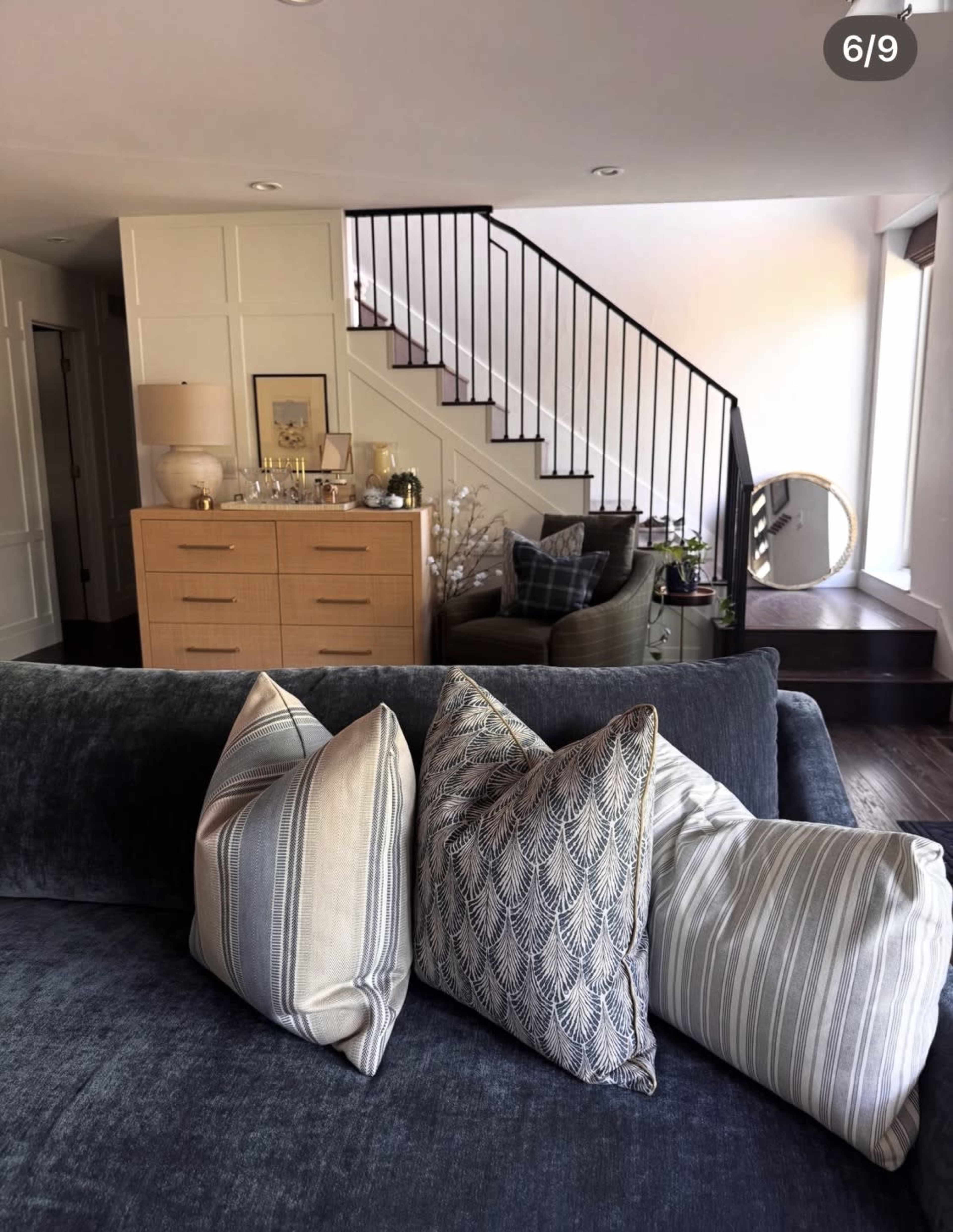 The image shows a modern living room with a dark sofa adorned with decorative pillows, a wooden dresser against the wall, and a staircase leading to an upper level.