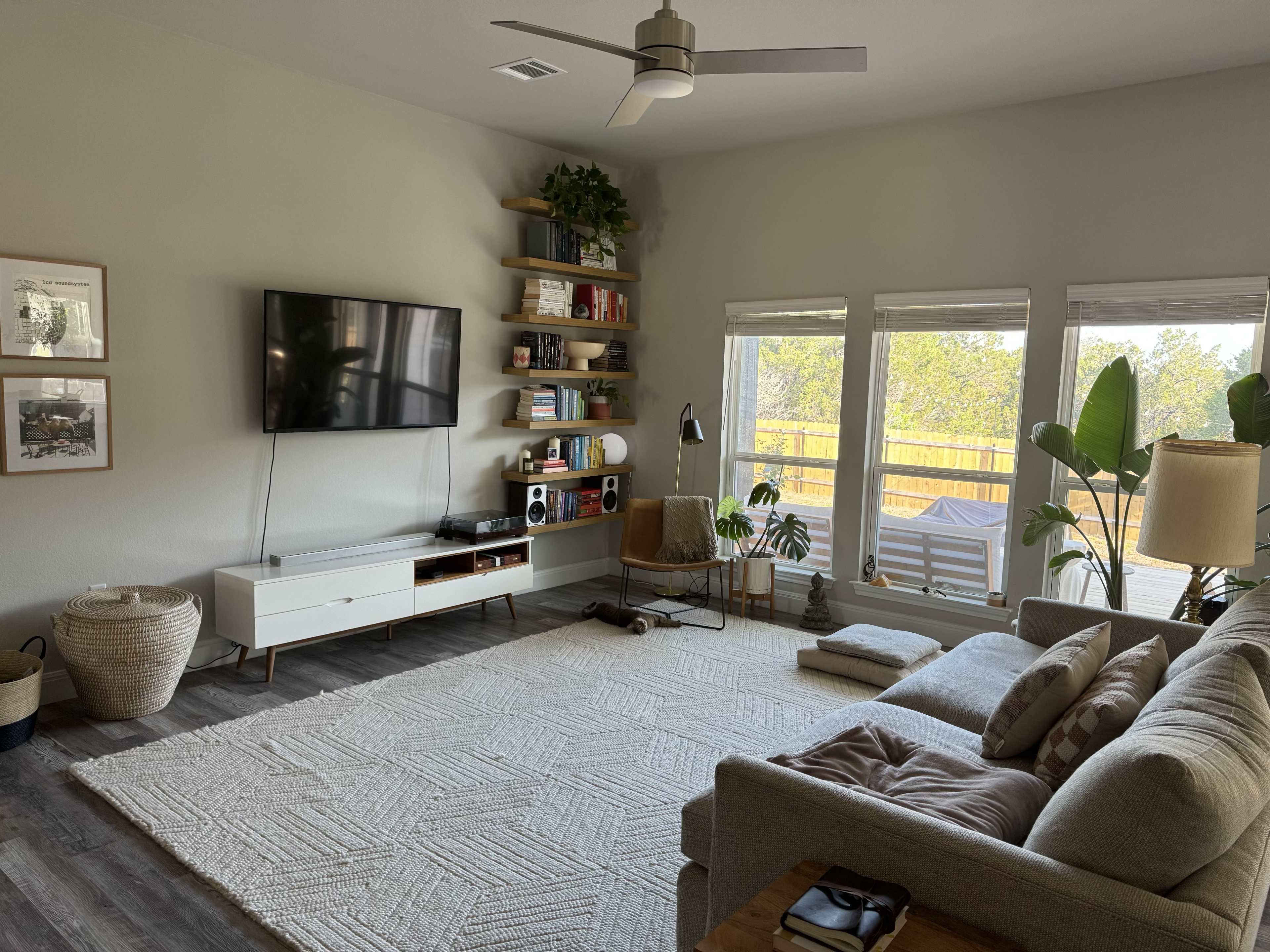 A bright living room features a large window, a sectional sofa, a television mounted on the wall, and a bookshelf filled with books and decorative items.