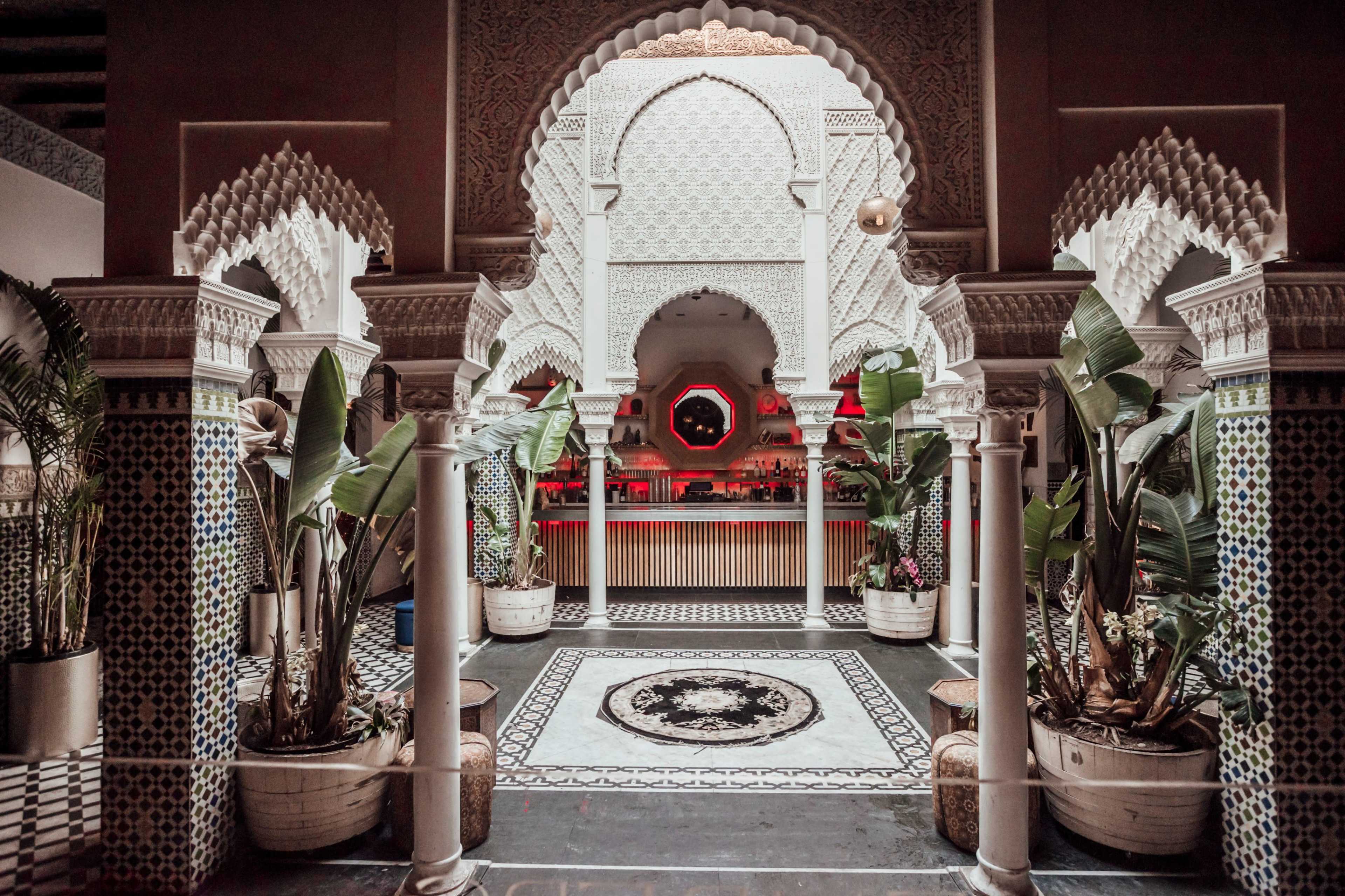 An intricately designed Moroccan-style courtyard with decorative tilework, arched doorways, and lush greenery in large planters.