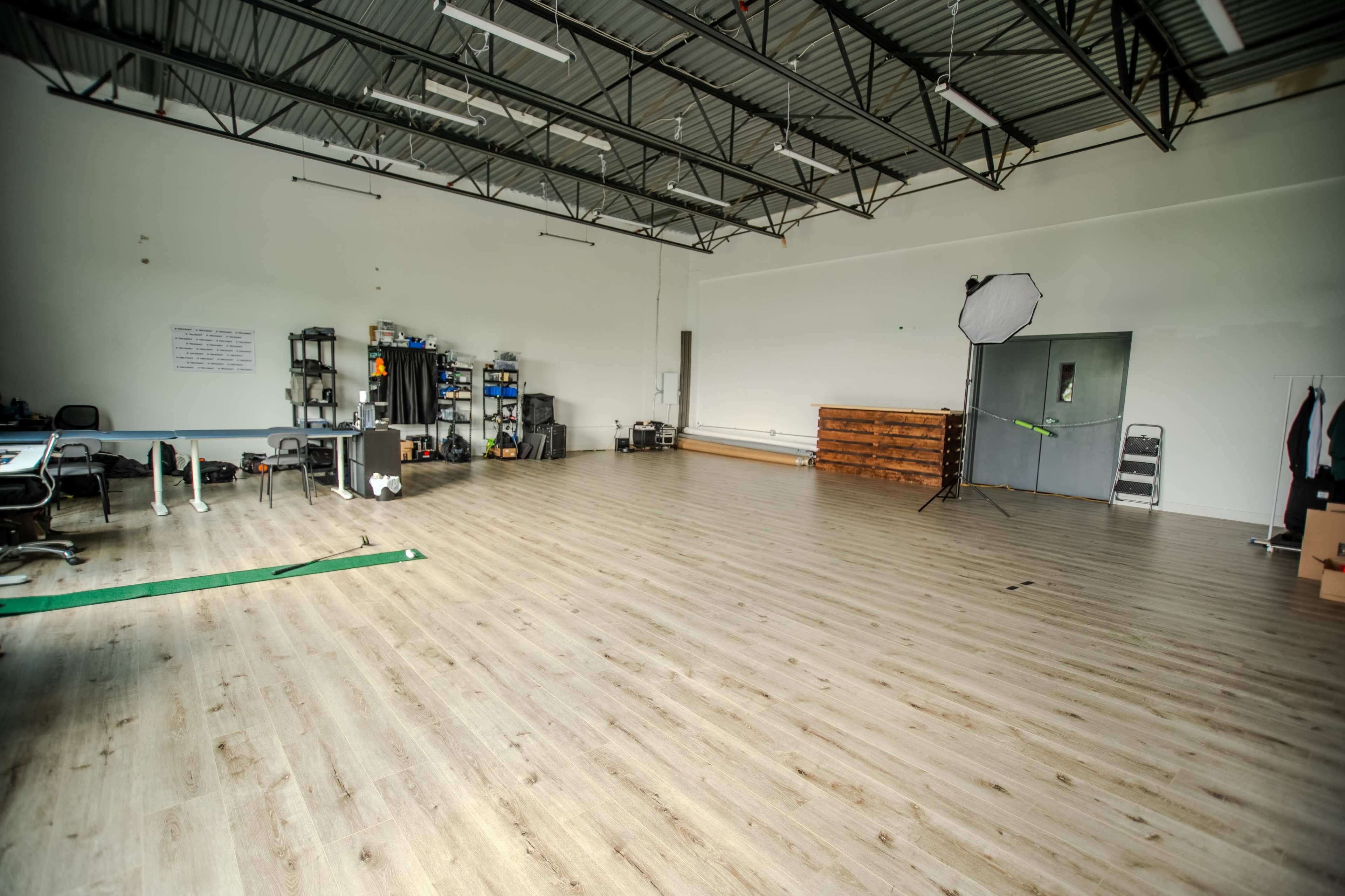 The image shows a spacious, empty studio with wooden flooring, various equipment along one wall, and a large backdrop setup in the corner.