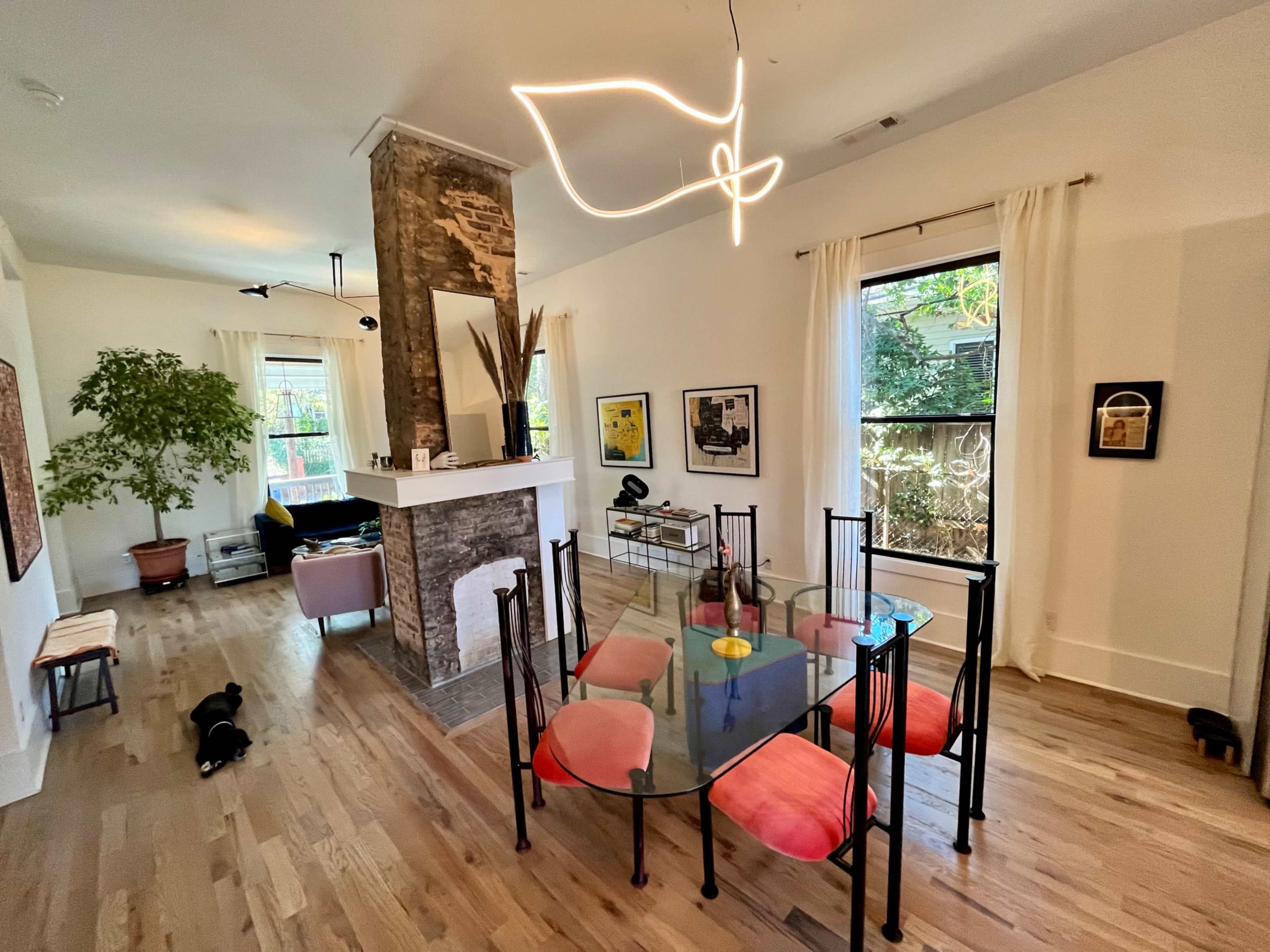 A dining area features a glass table surrounded by black chairs, with a modern light fixture above and a stone fireplace in the background.
