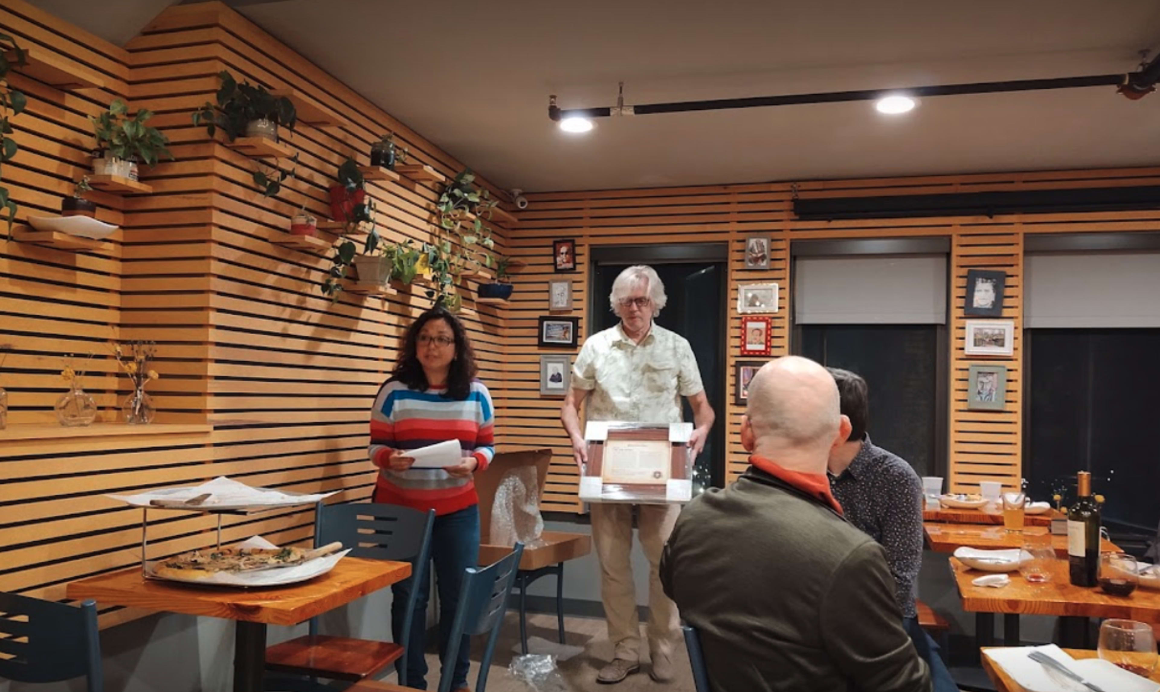 Two individuals are standing in a restaurant with wooden paneling; one is holding a box while the other is holding papers, and a few patrons are seated at tables in the foreground.