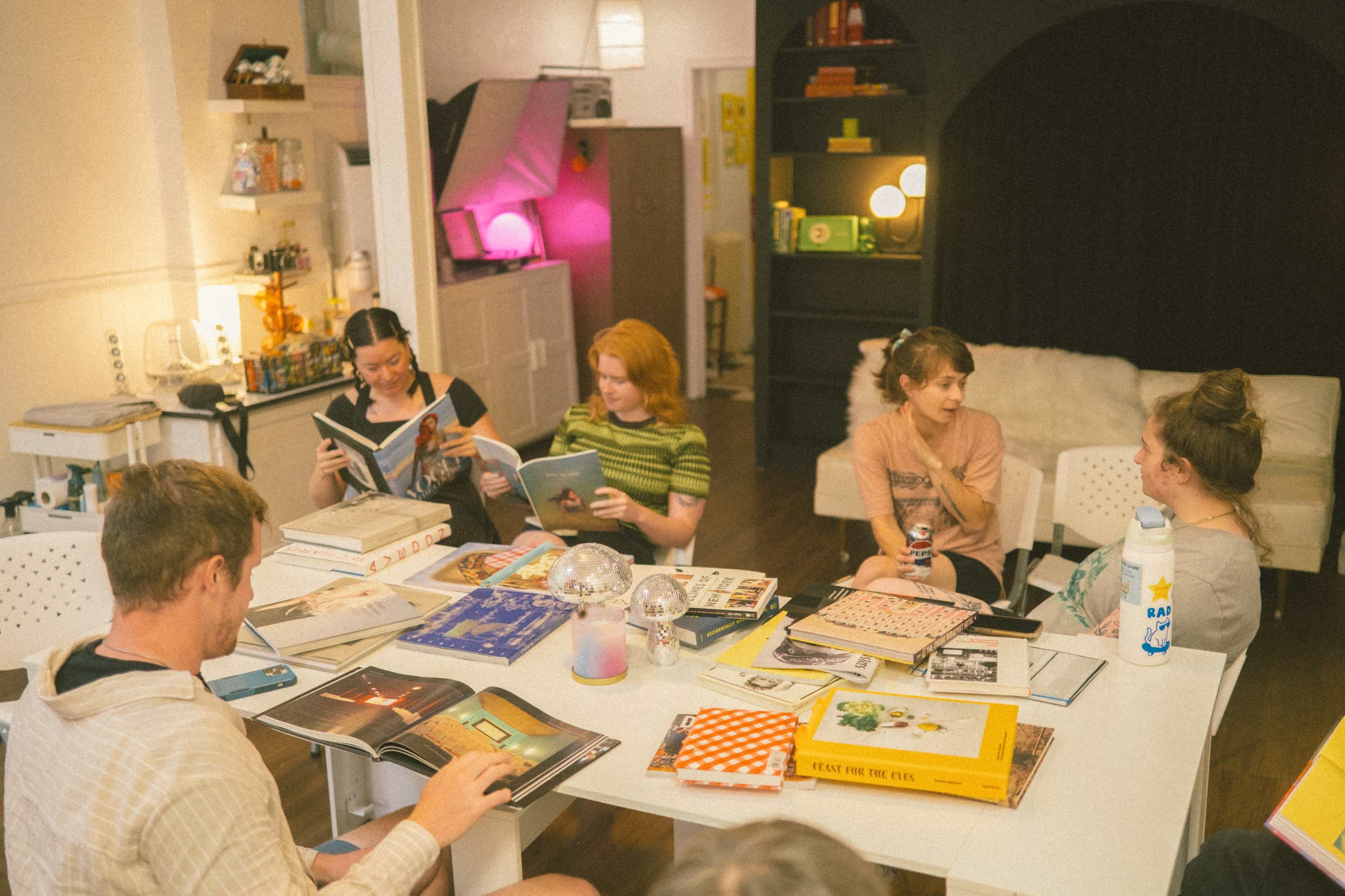 A group of people sits around a table in a well-lit room, engaging with various magazines and books.