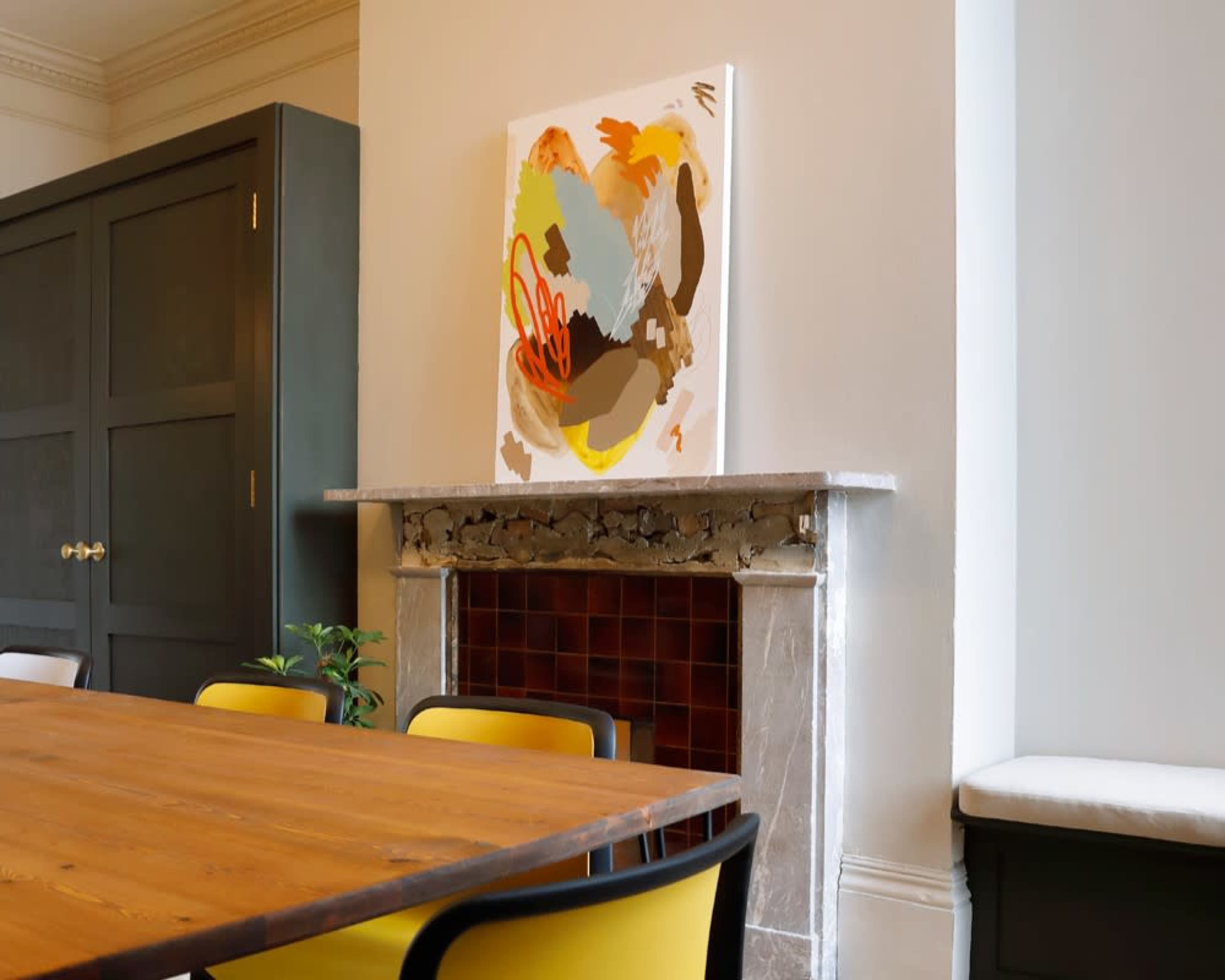 The image shows a dining area featuring a wooden table surrounded by yellow and black chairs, with a colorful abstract painting displayed above a stone fireplace.