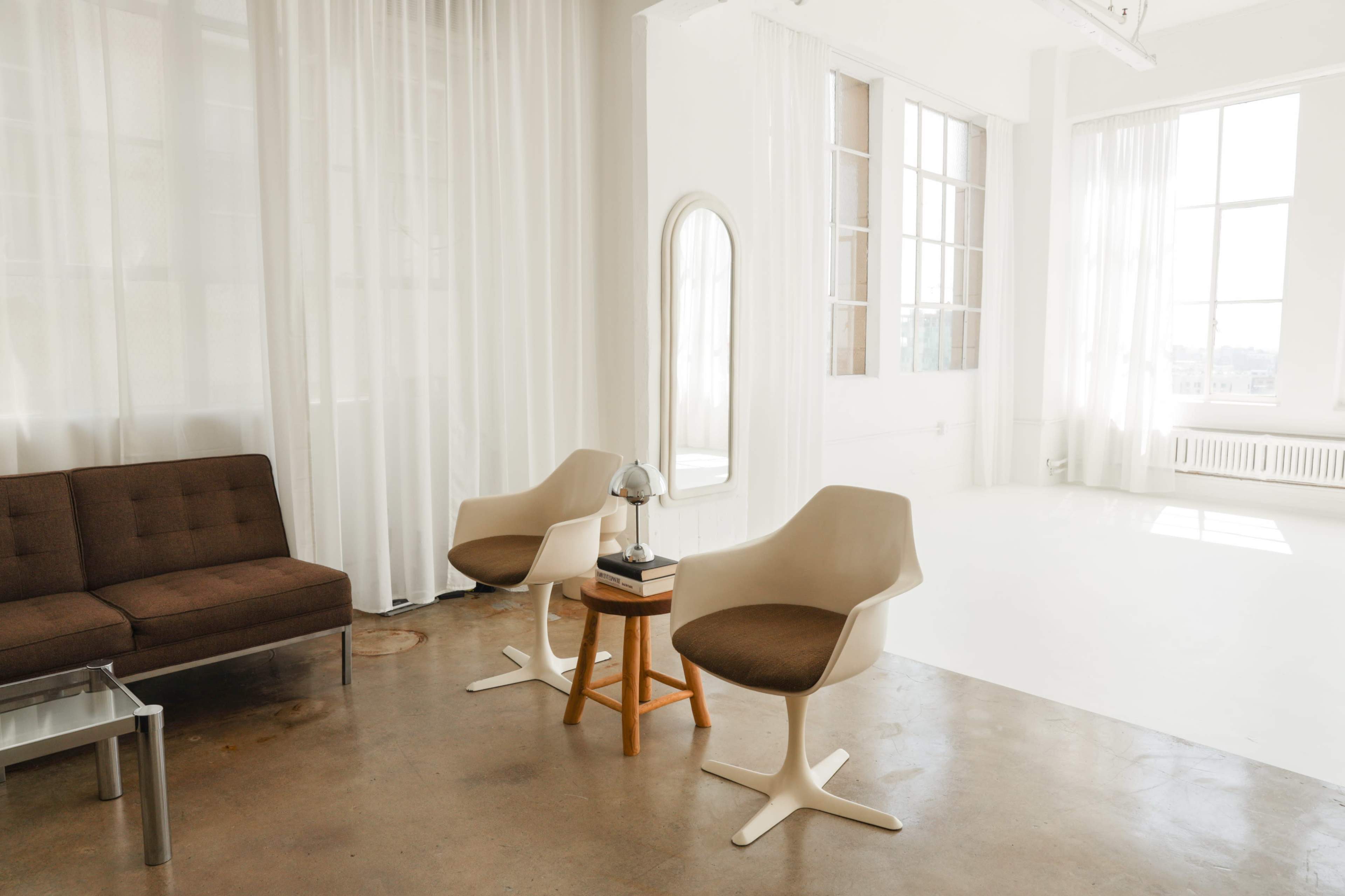 A bright, minimalistic interior with a brown sofa, two white chairs, a small table with a lamp, and large windows covered with sheer curtains.