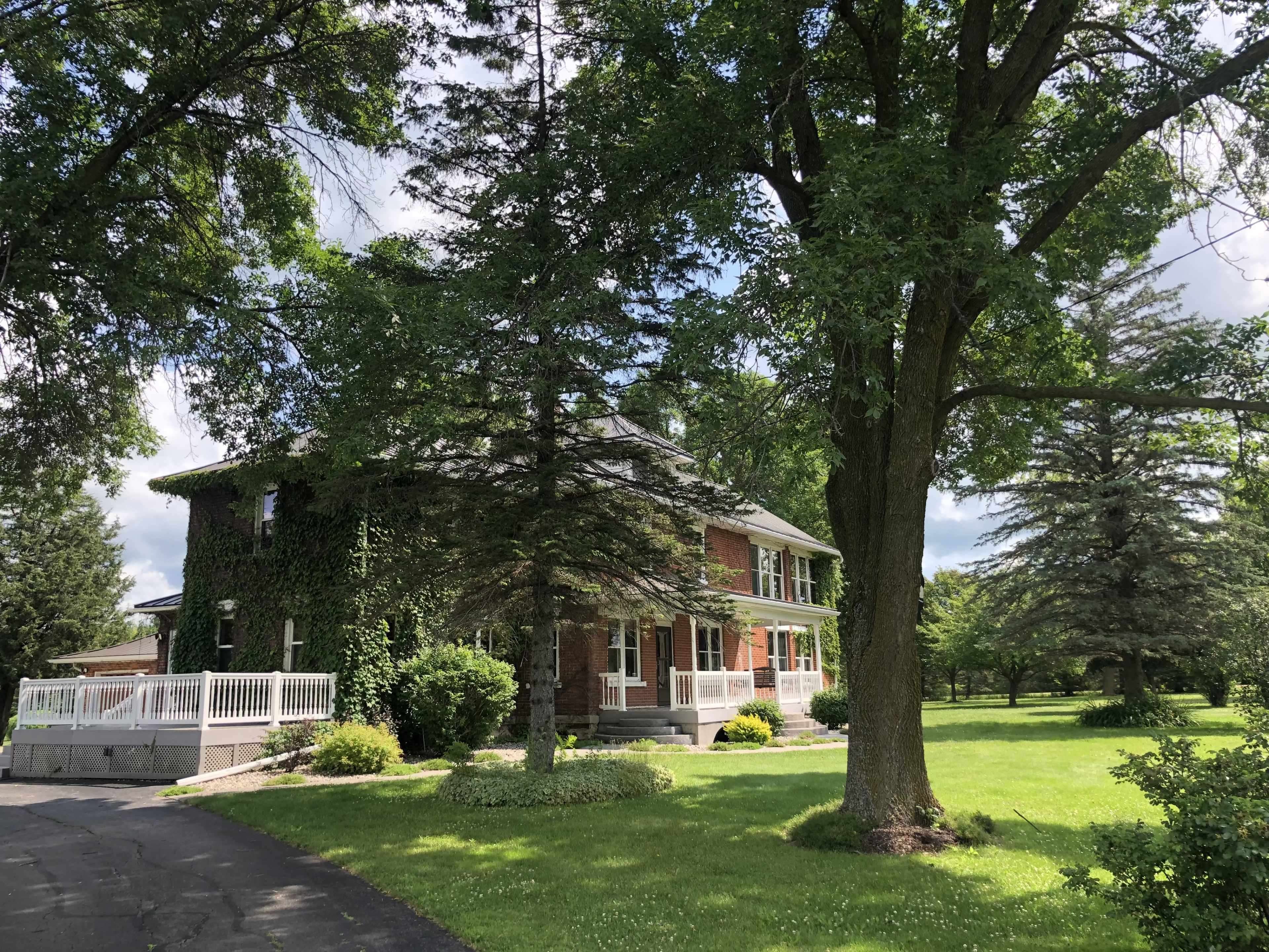 A brick house covered in ivy sits beside a large tree and manicured lawn under a partly cloudy sky.