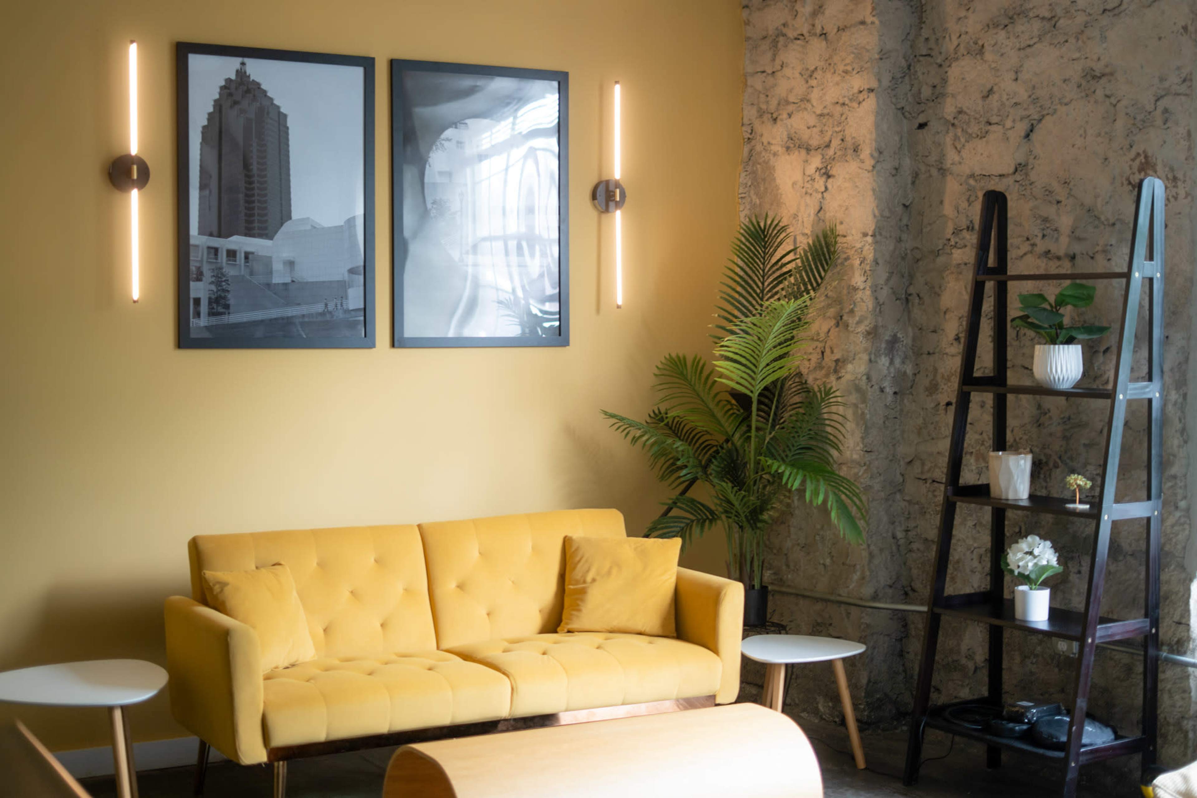 The image shows a cozy interior featuring a yellow sofa, two black and white framed pictures on the wall, a potted plant, and a wooden ladder shelf against a textured wall.