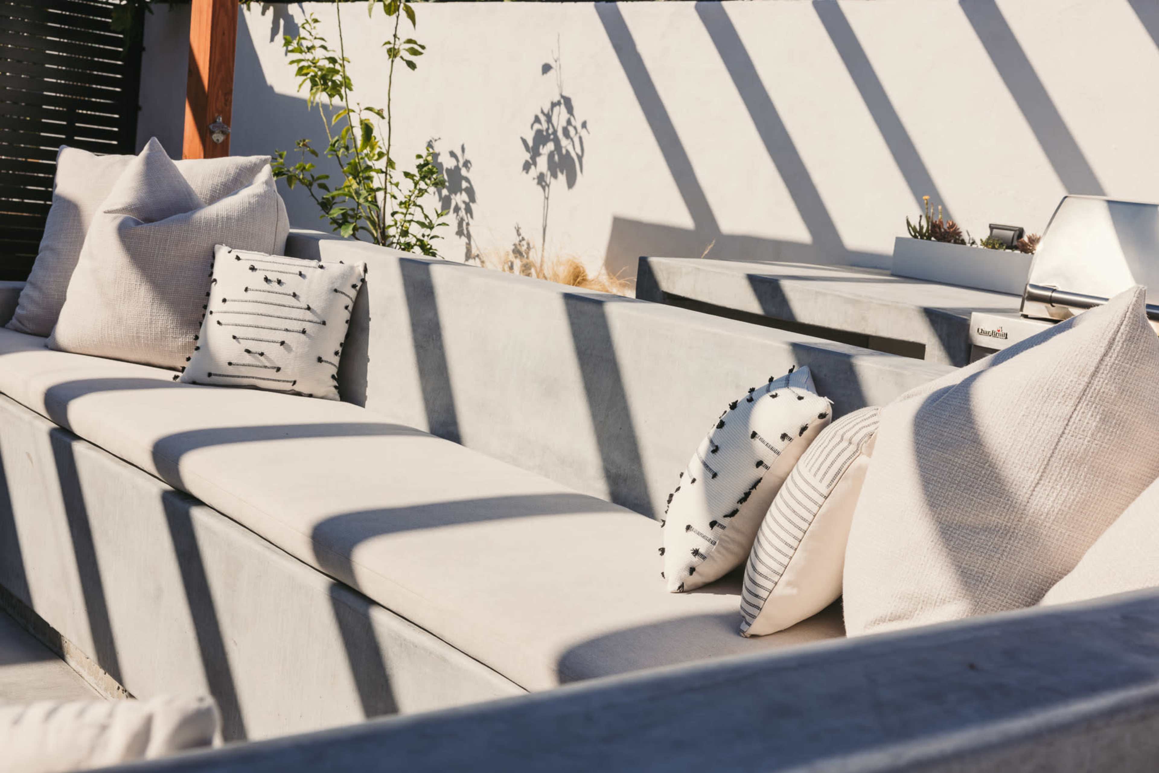 A concrete seating area with cushions and decorative pillows, illuminated by shadows from nearby structures.