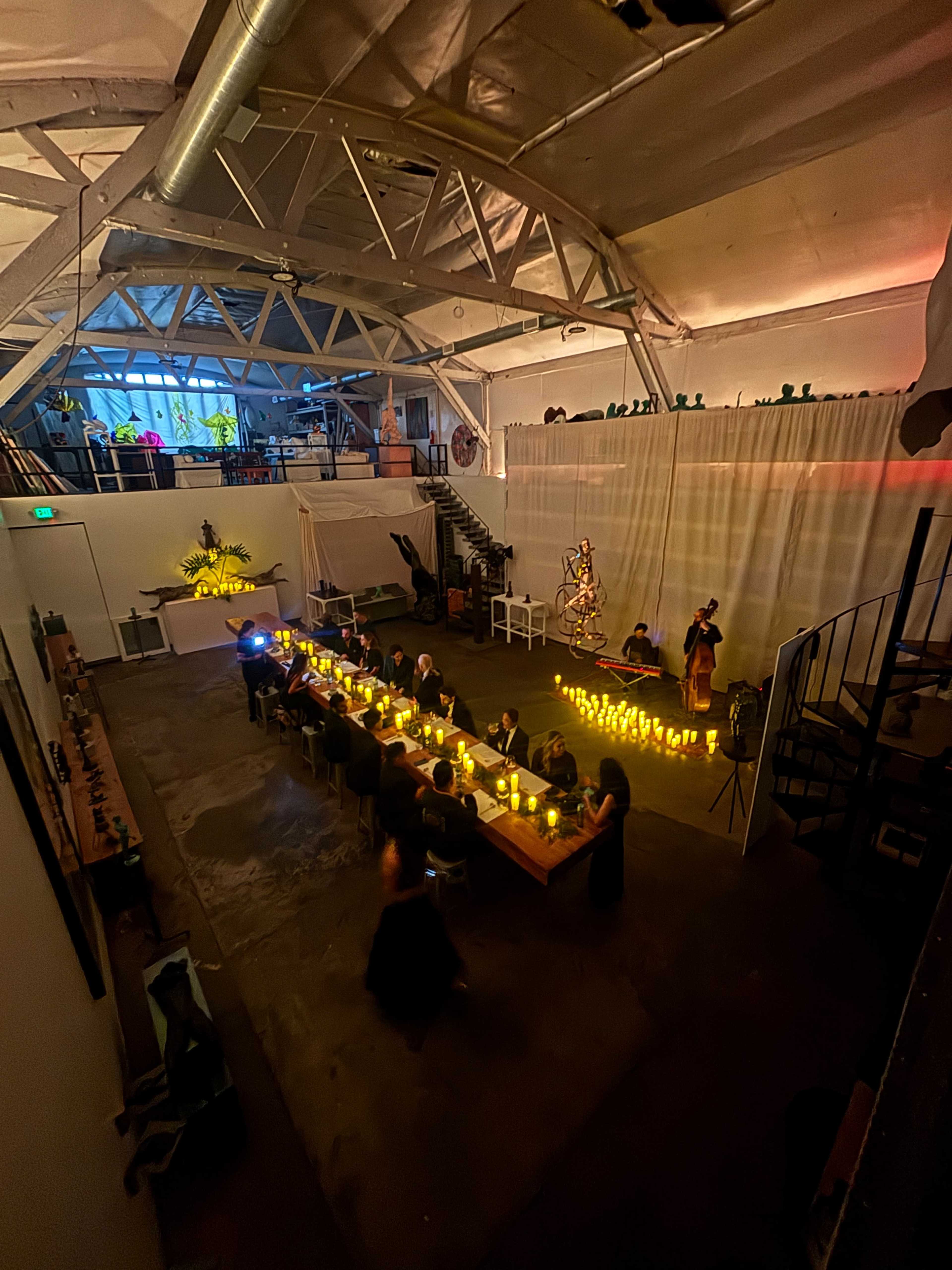 A long table with candles is set up in a large, industrial-style venue, where guests are seated and engaged in conversation amidst decorative lighting and plants.