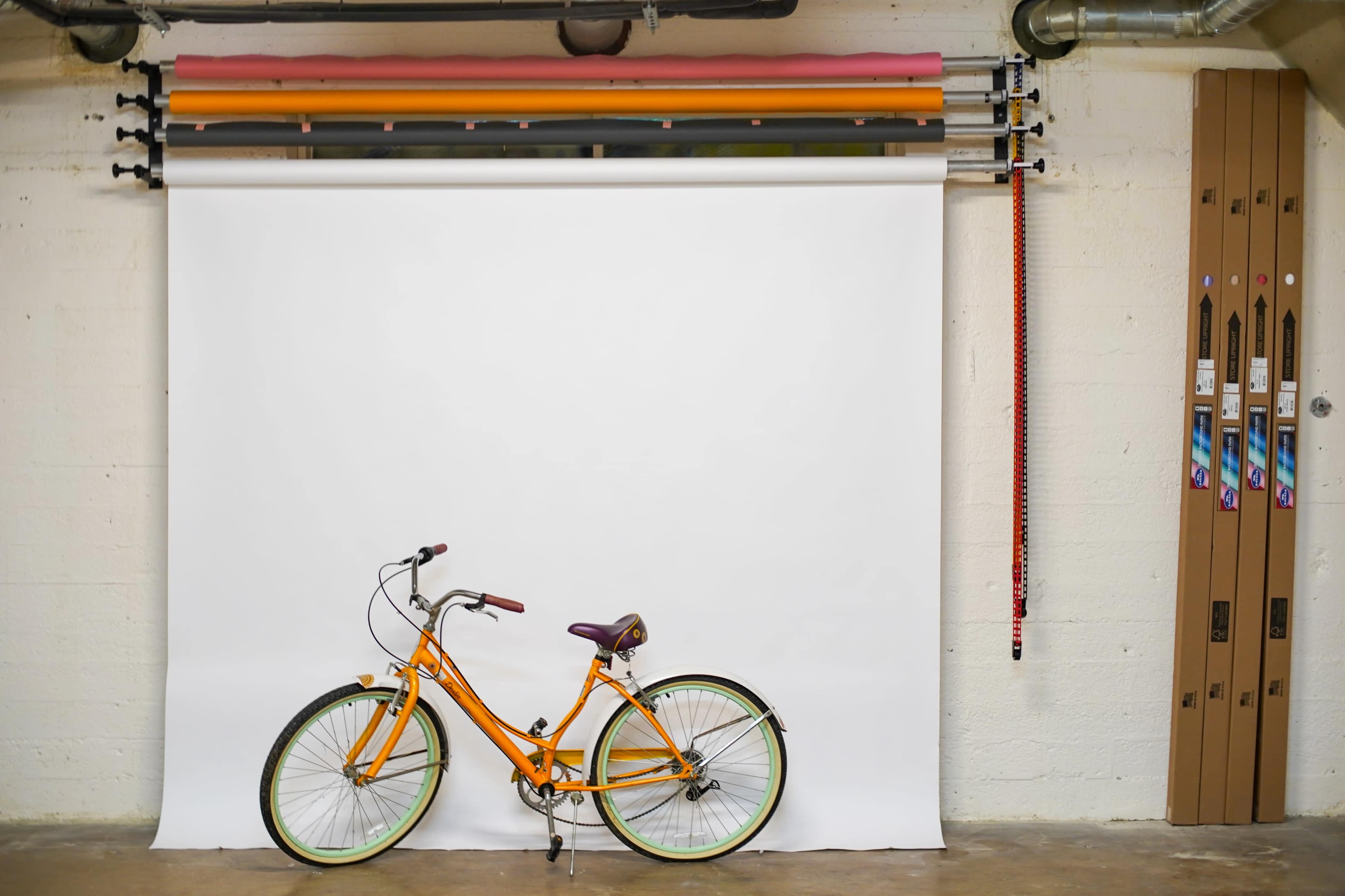 An orange bicycle is parked on a concrete floor beside a white backdrop in a basement space.
