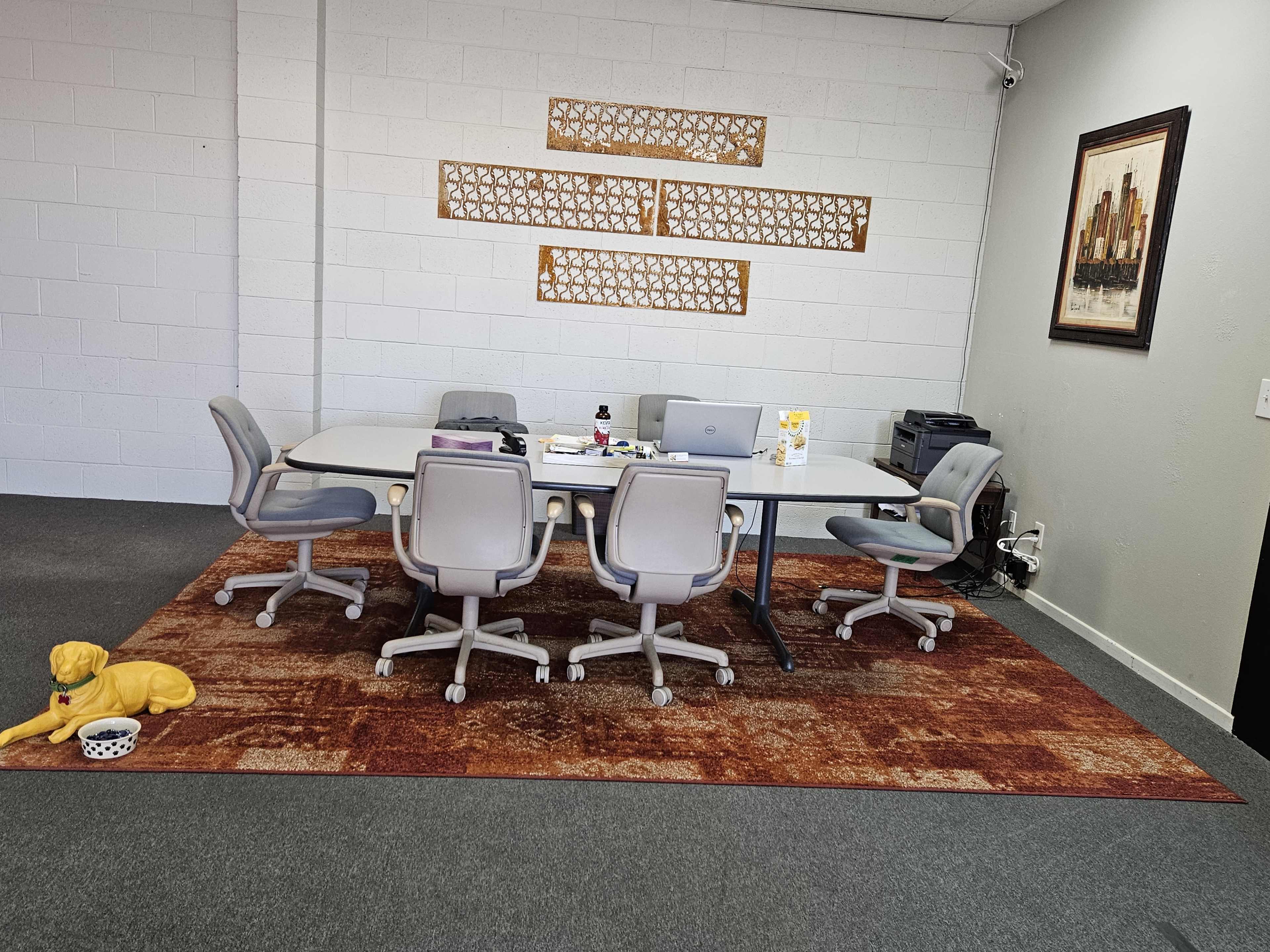 A meeting room features a large table surrounded by six gray chairs, a computer, and a decorative rug, with a dog lying on the floor beside a bowl.