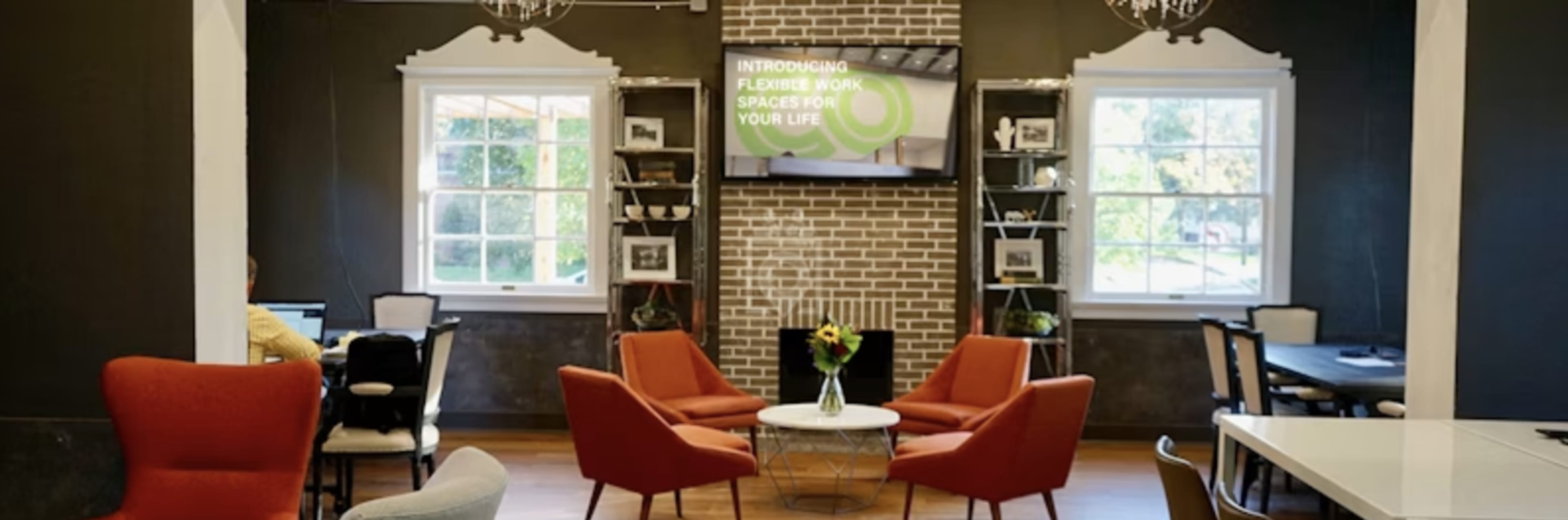 The image features a modern workspace with red chairs, a central table, a brick accent wall, and large windows allowing natural light.
