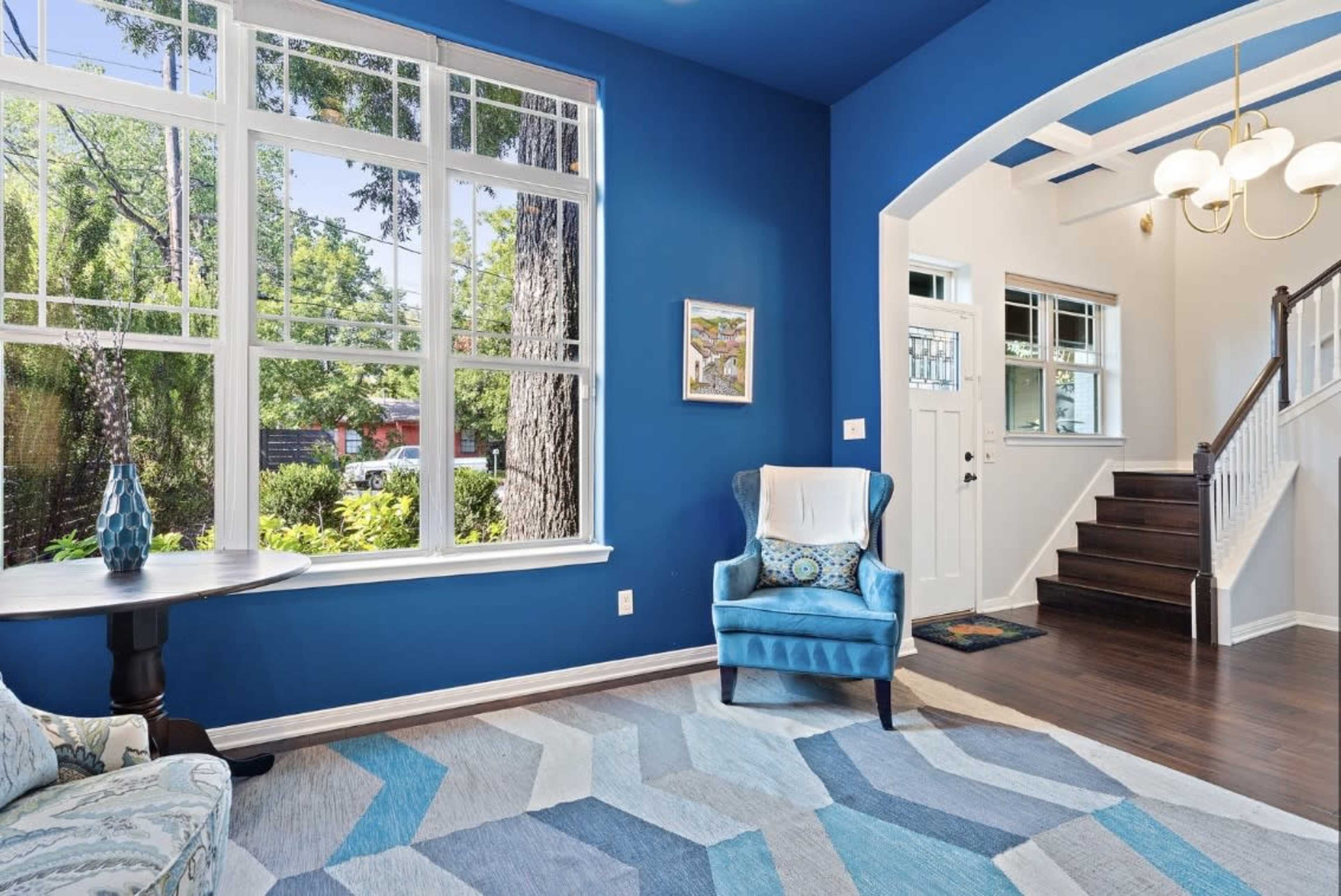 A bright living room features a blue accent wall, large windows overlooking greenery, and a staircase leading to the upper floor.