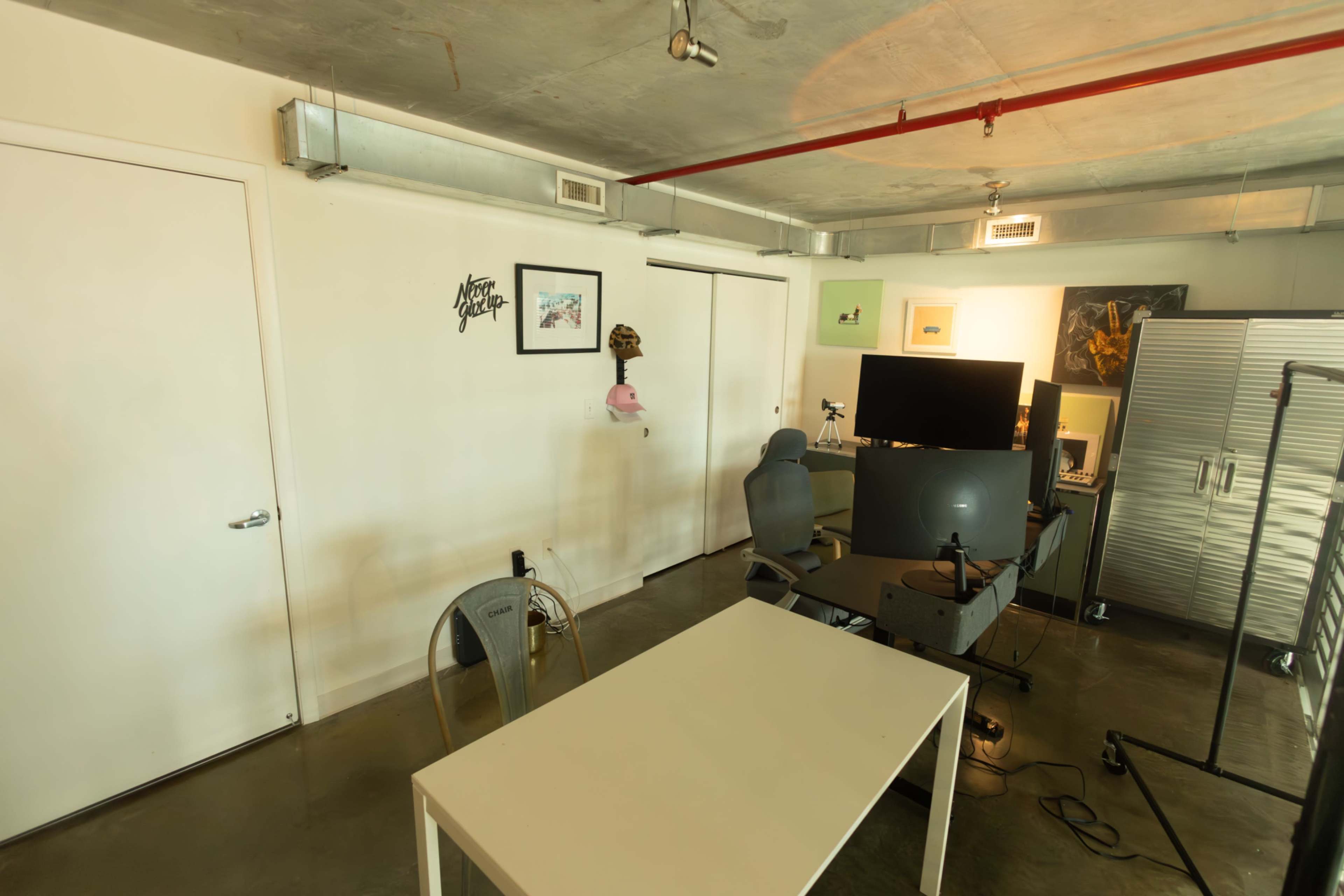 The image shows a modern office space with a white table, two gray chairs, a computer desk with a monitor, and various wall decorations.