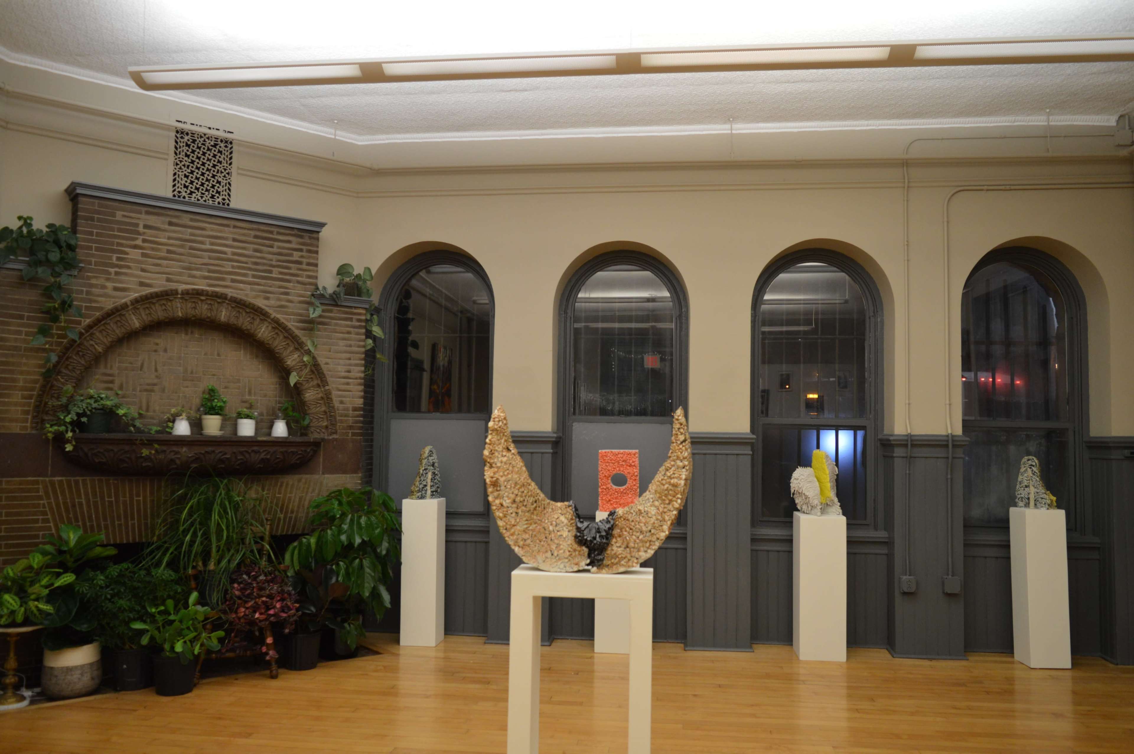 An indoor exhibition space with several sculptures displayed on pedestals, surrounded by greenery and arched windows.