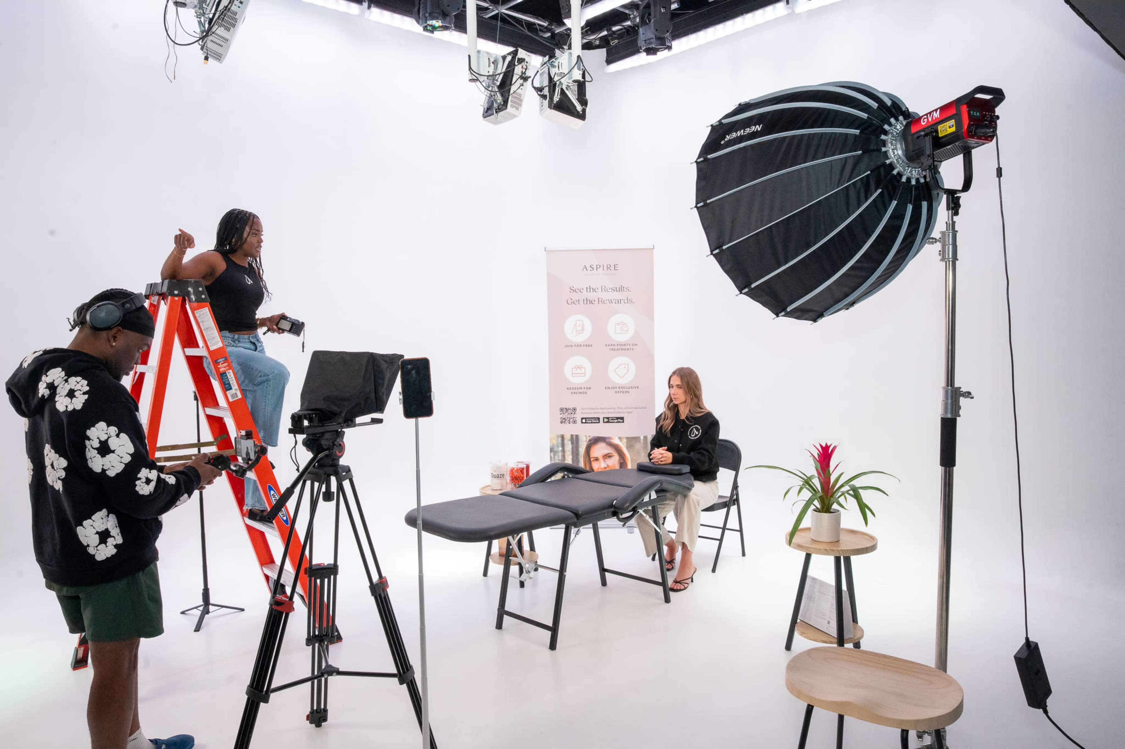 A filming set is shown with a woman on a ladder adjusting lights, two chairs, a table, and various equipment arranged on a white backdrop.