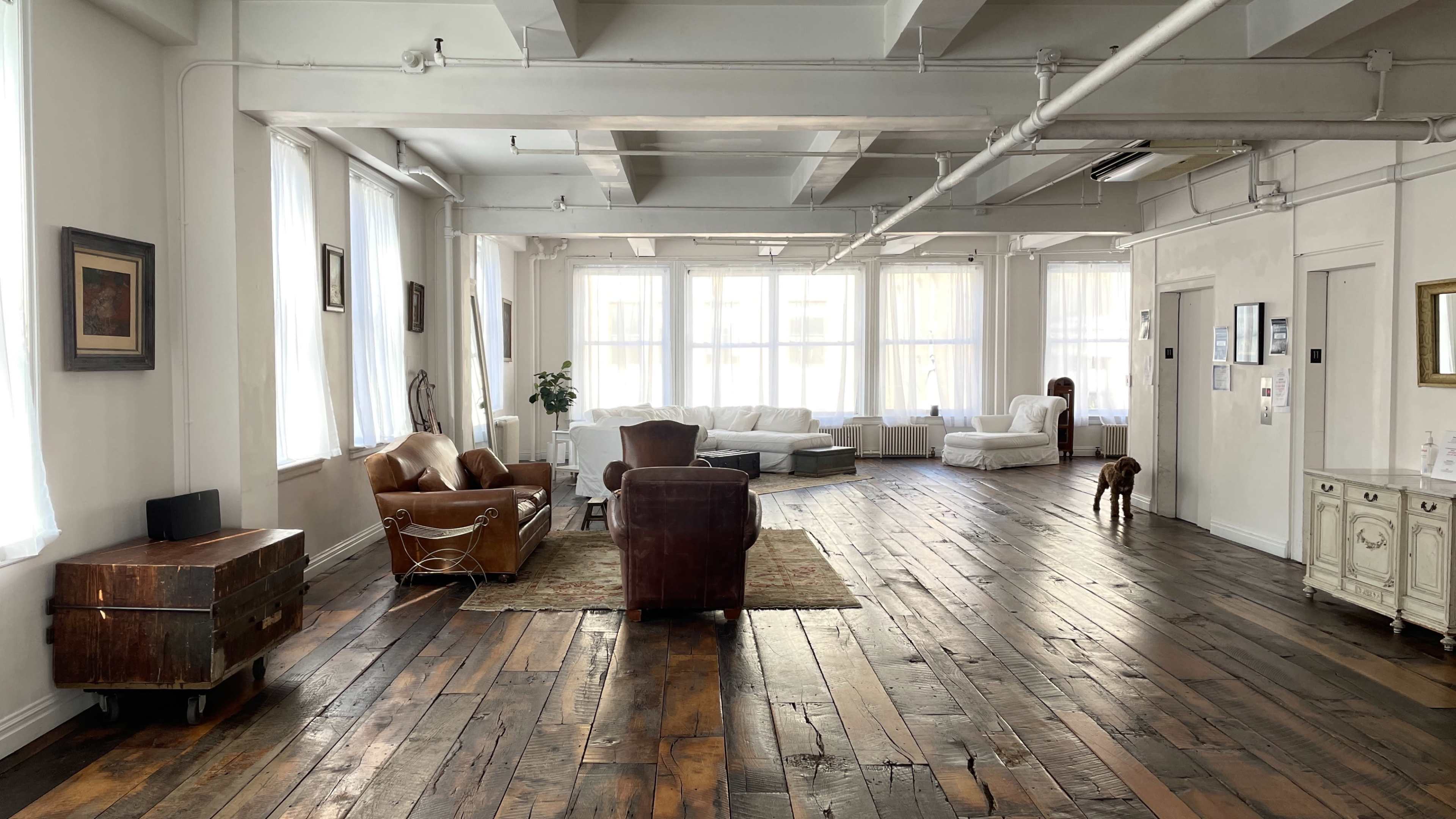 A spacious, bright room with wooden floors, large windows, and a mix of seating areas, including a couch and armchairs.