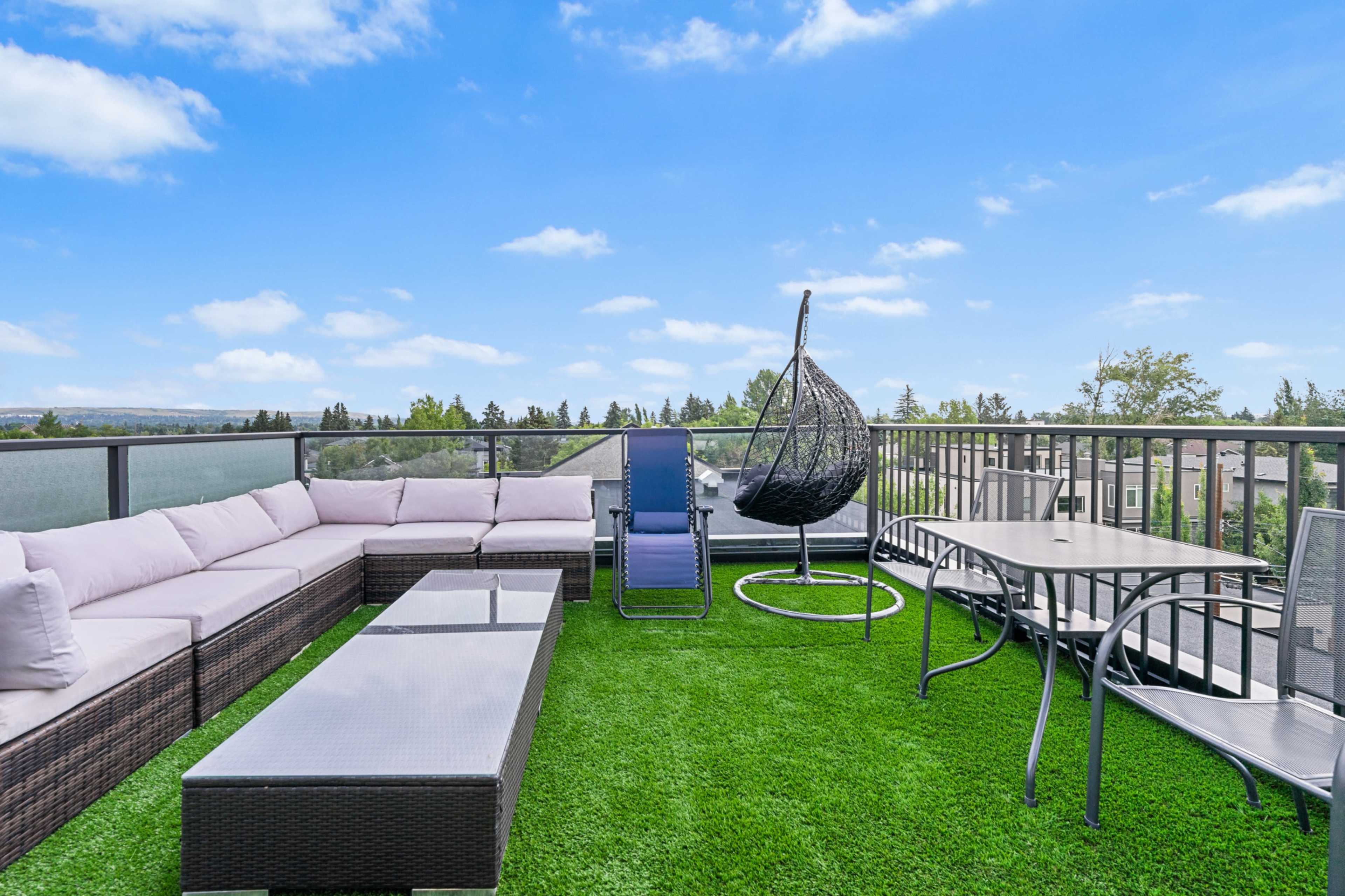 A rooftop terrace features a wicker sectional sofa, a hanging chair, a lounge chair, and a dining table, all set on green artificial grass.