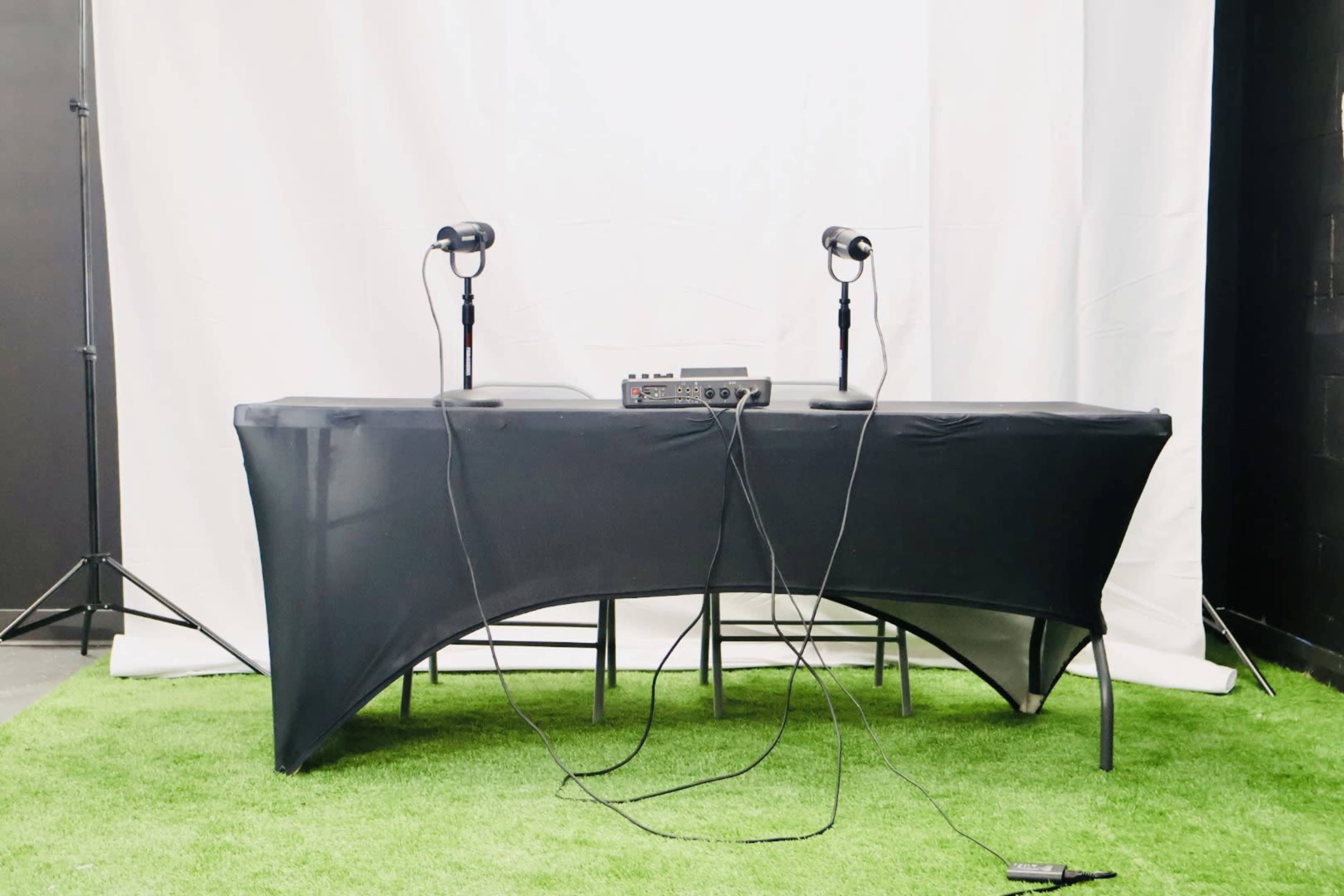 A black table with two microphones and an audio mixer is set against a white backdrop and green artificial grass.