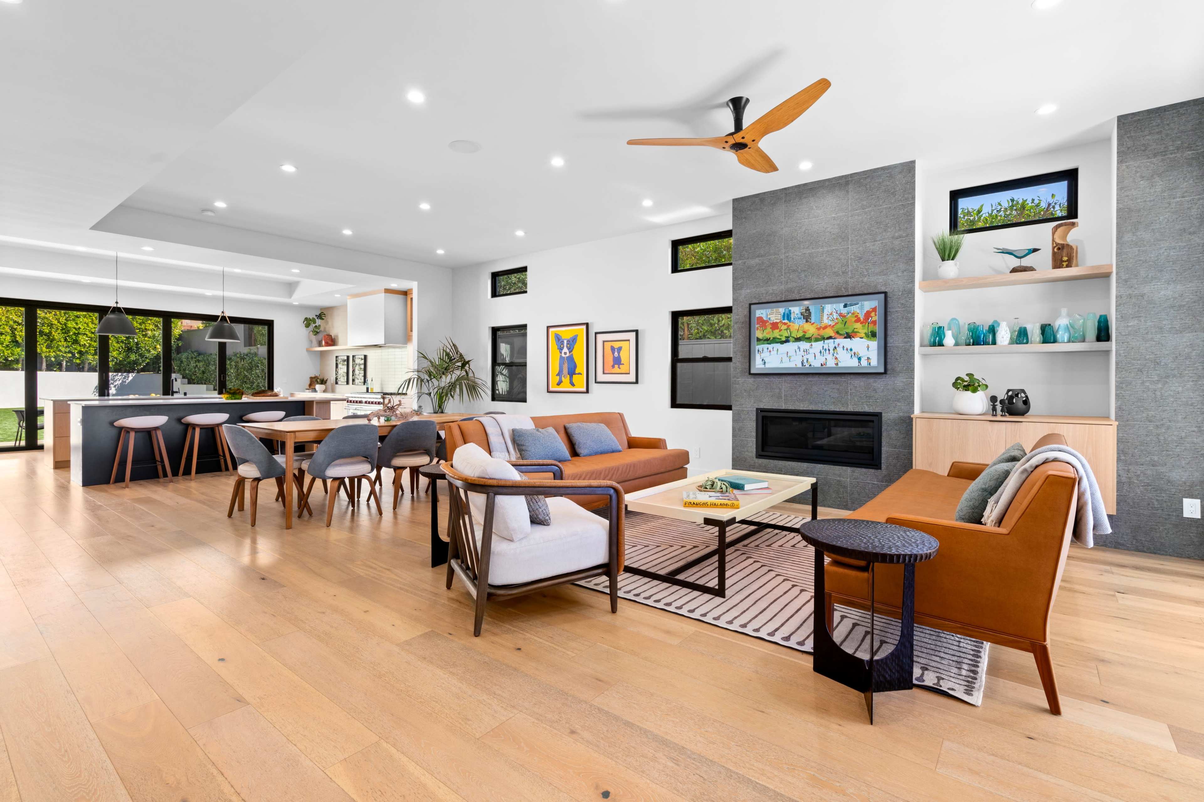 A modern, open-concept living area with a seating arrangement, a television, a kitchen bar, and large windows overlooking greenery.